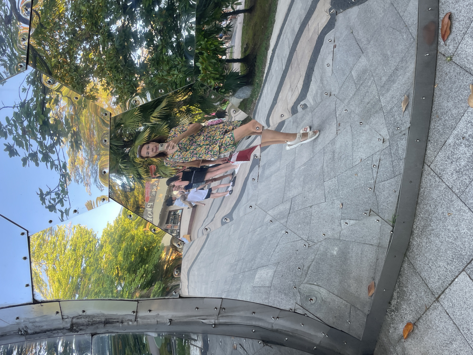 A woman is taking a selfie in a mirrored sculpture in Sochi, Russia. The sculpture is made of hexagonal mirrored panels and is located outdoors, possibly in a park or public space, as indicated by the presence of trees, greenery, and a tiled walkway. The woman is wearing a floral print dress and white sandals. In the reflection of the sculpture, other people can be seen, including children, walking in the background. The sculpture distorts the reflections, creating a wavy, fragmented image of the surroundings. The ground is paved with large grey tiles, and some dry leaves are scattered on the ground.