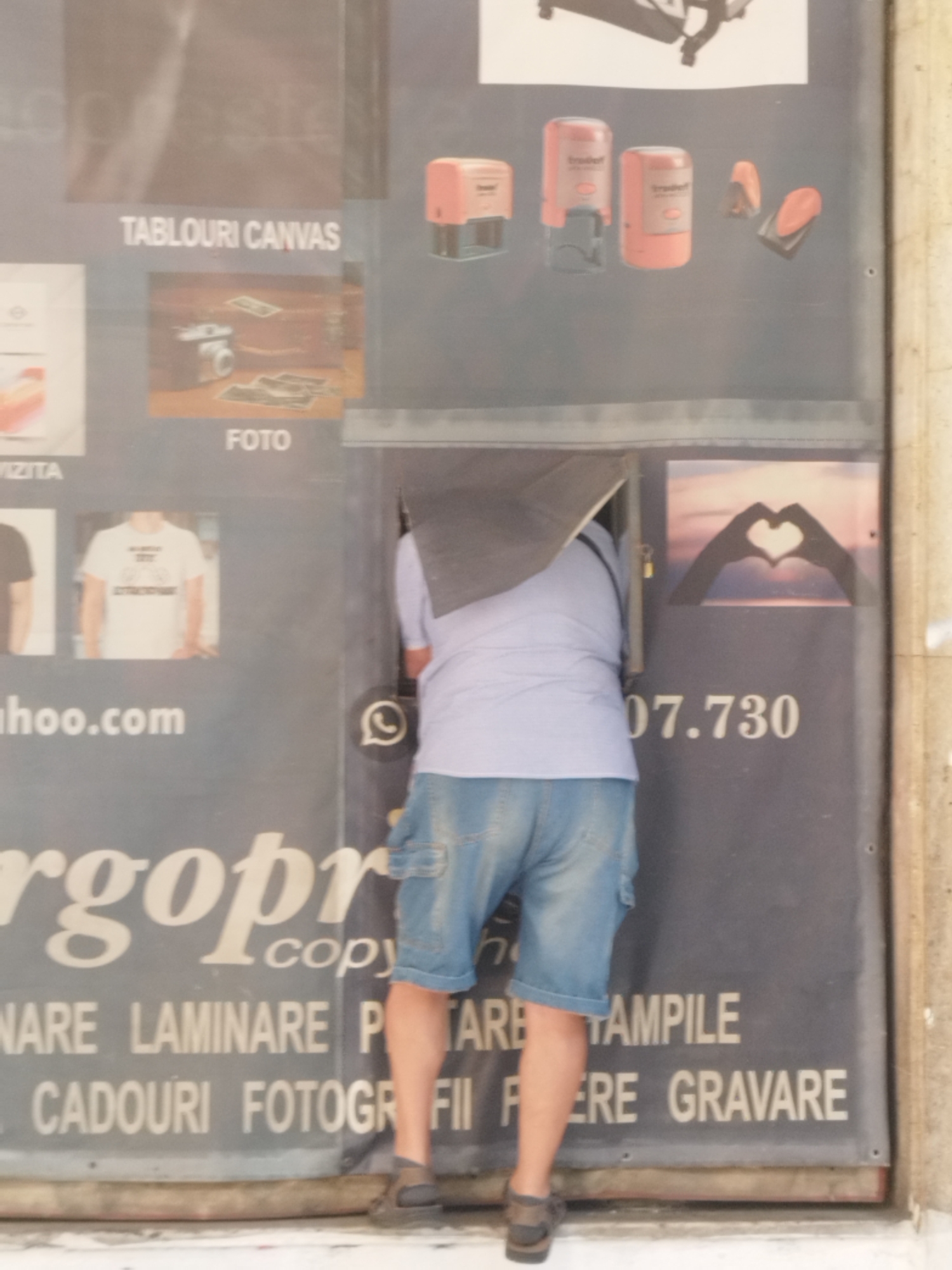A person wearing denim shorts and a light blue shirt is leaning into an opening in a dark-colored advertisement board. The opening appears to be a slot or a small window. The advertisement board displays text and images, including "TABLOURI CANVAS", "FOTO", a camera, and a heart shape formed by hands. There is also a phone number, "07.730", visible on the board. A WhatsApp icon is also present. The lower portion of the advertisement features text such as "NINARE LAMINARE P TARE", "CADOURI FOTOGR FII ERE GRAVARE". The scene appears to be outdoors, possibly on a street in Bucharest, Romania, given the Romanian text on the advertisement.