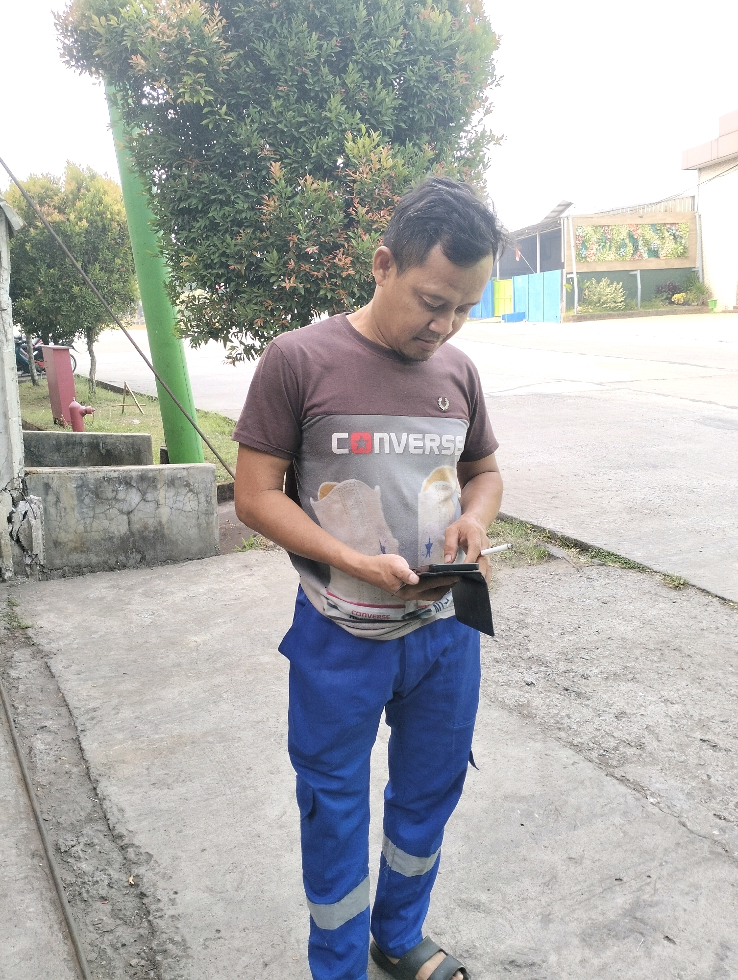 A middle-aged East Asian man is standing outdoors in a paved area near some trees. He is wearing a brown t-shirt with a Converse logo and an image of a shoe, and blue pants with reflective stripes. He is looking down at a black wallet in his hands. In the background, there are trees, a green pole, and buildings. The image appears to be taken in Gunung Sindur, Indonesia.