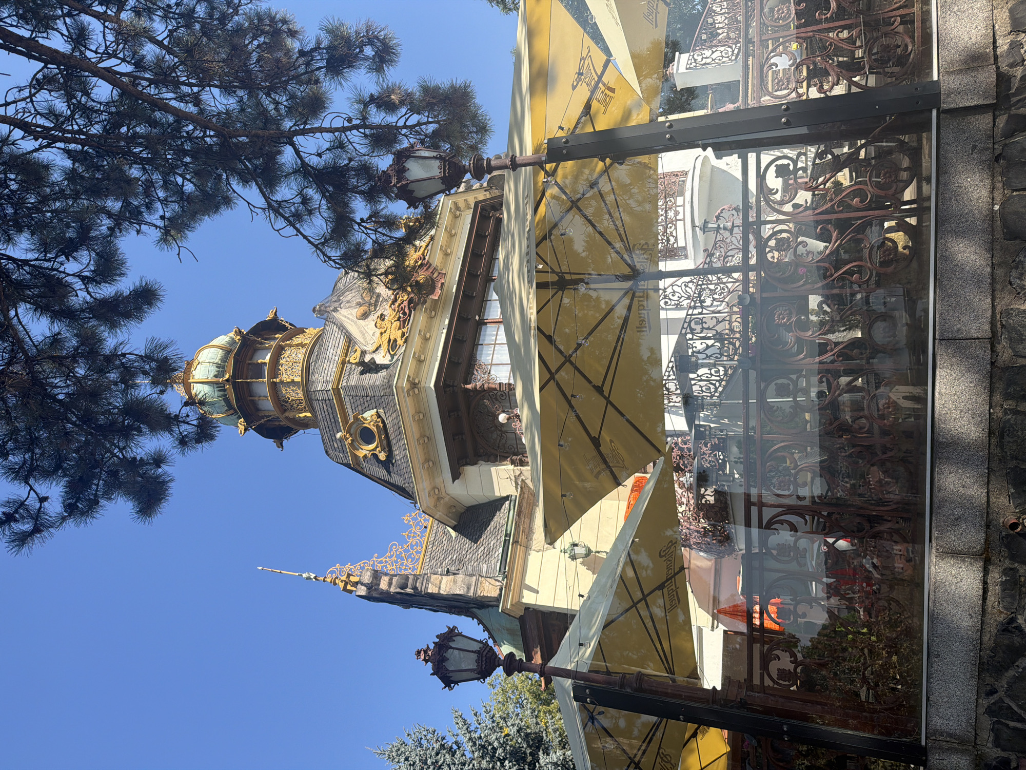 A bright, sunny day showcases the ornate architecture of a building in Prague, Czech Republic, likely a historic establishment given its elaborate design. The building features a prominent golden dome topped with a smaller green dome, intricate gold detailing on its facade, and a grey tiled roof. In the foreground, several large, yellow umbrellas are open, suggesting an outdoor seating area for a cafe or restaurant. A rustic, dark metal lamppost stands to the left of the frame. The scene is framed by the dark green branches of a pine tree in the upper right corner, casting shadows onto the building and umbrellas. A stone wall forms the base of the foreground, with a decorative metal railing visible behind the umbrellas, reflecting the surroundings in the glass. The sky is a clear, vibrant blue.