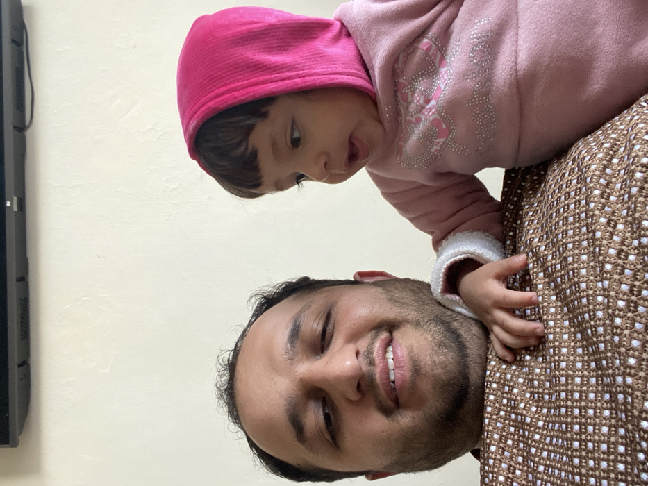 The image displays an adult male and a young child indoors in Kandahar, Afghanistan. The adult male, positioned on the left side of the frame, has dark hair and a short beard, smiling slightly towards the camera. He wears a brown garment featuring a textured, white, diamond-patterned weave. To his right, a young child, an infant or toddler, is partially visible, wearing a pink hooded, long-sleeved outfit with white trim on the left cuff and silver and pink decorative elements on the chest. The child's left hand rests on the adult male's right shoulder. The child is looking towards the right side of the frame with a slightly open mouth. The background consists of a plain light-colored wall, above which a black, rectangular electronic device is mounted horizontally.
