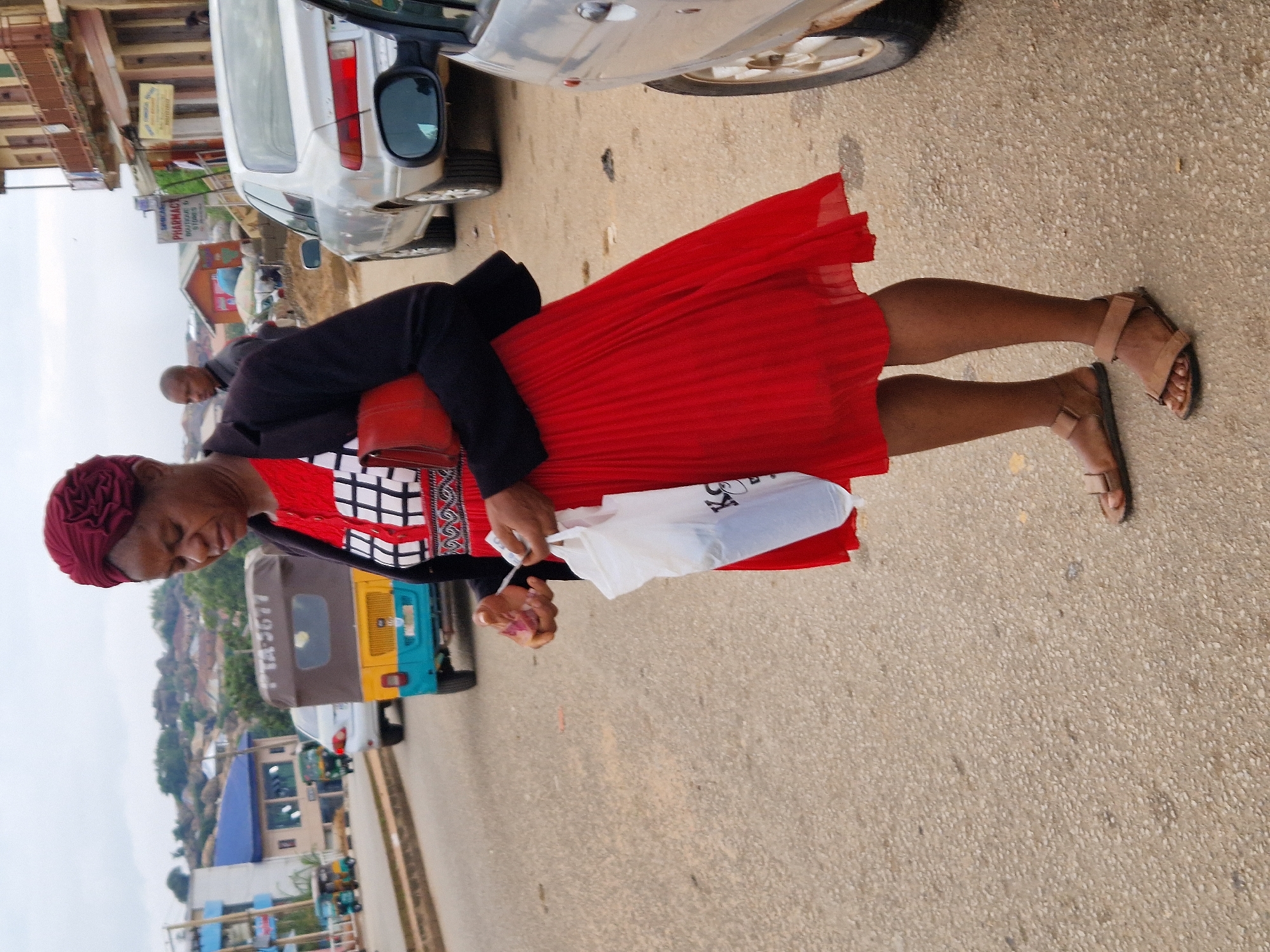 In an outdoor, urban setting, a woman stands on a paved road, looking downwards and holding a white plastic bag and a red clutch in her hands. She is wearing a red headscarf, a black jacket over a red and white patterned top, and a flowing red pleated skirt. Her feet are clad in brown sandals. Behind her, a yellow and blue auto-rickshaw is parked on the side of the road, and a silver sedan is driving past. In the background, there are buildings, some of which appear to be shops, and a hilly landscape with more structures visible. The sky is overcast.