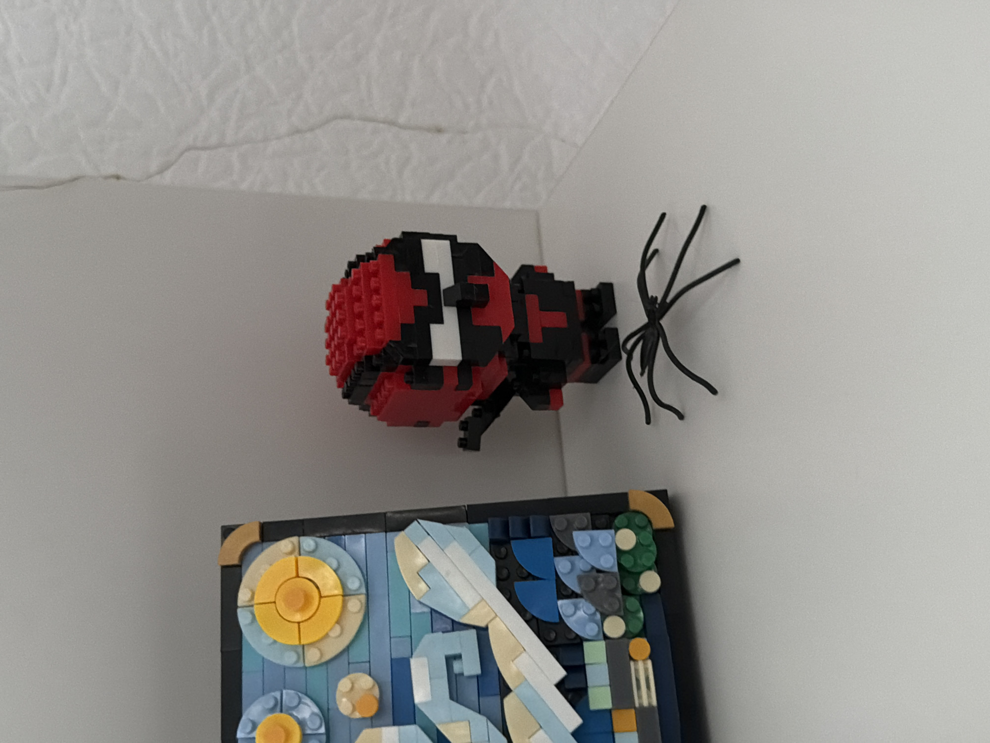 A close-up, eye-level shot captures a white surface with two LEGO creations and a small black spider figure. On the left, a framed LEGO mosaic depicts a stylized starry night sky with yellow and blue circles representing celestial bodies and swirling blue and white elements suggesting clouds or waves. The frame is black with a tan corner piece. To the right of the mosaic stands a LEGO minifigure resembling a cartoon character, primarily red and black with white eye details and a black "T" on its chest. In front of the minifigure, a black, four-legged spider figure is positioned. The background consists of a textured white surface on the right, possibly a wall, and a plain grey area above the LEGO creations. There are no visible people or any indication of the city of Kyiv, Ukraine.