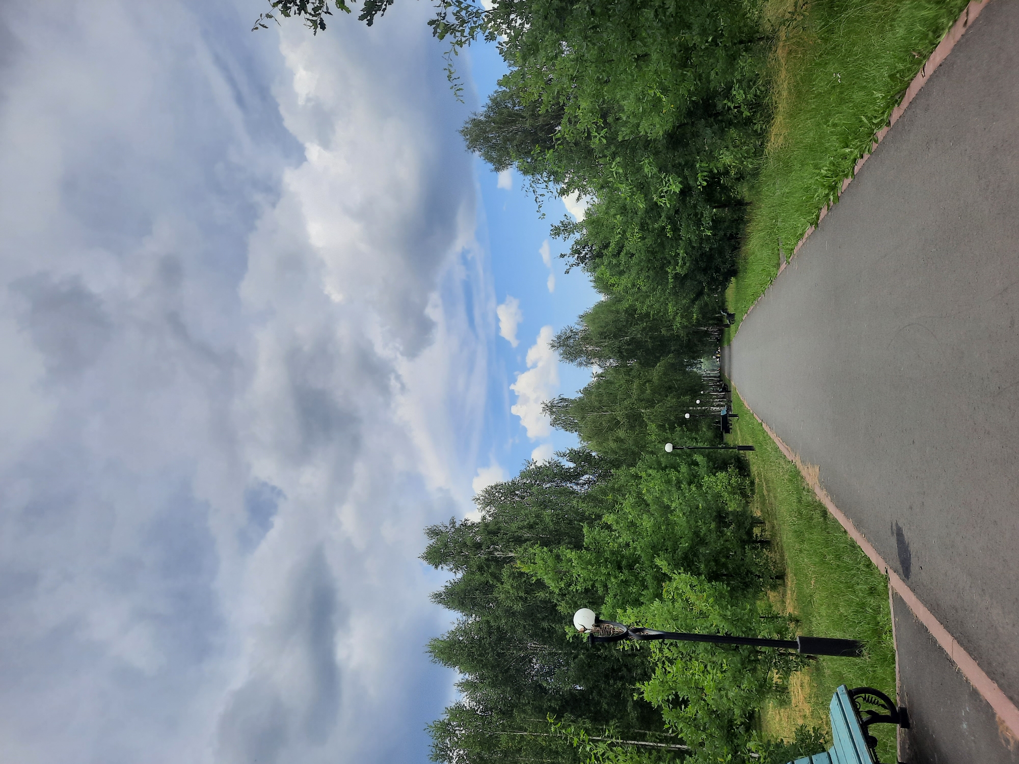 The image depicts a park pathway in the city of Zelenogorsk, Russia. A paved asphalt path stretches into the distance, bordered by lush green grass and trees. Several lampposts with spherical lights are visible along the path, indicating a public park setting. A blue park bench is situated on the left side of the path near the foreground. The sky above is filled with dramatic, grey clouds interspersed with patches of blue sky, suggesting an overcast day with the possibility of rain. The overall scene conveys a sense of tranquility and natural beauty within an urban environment.