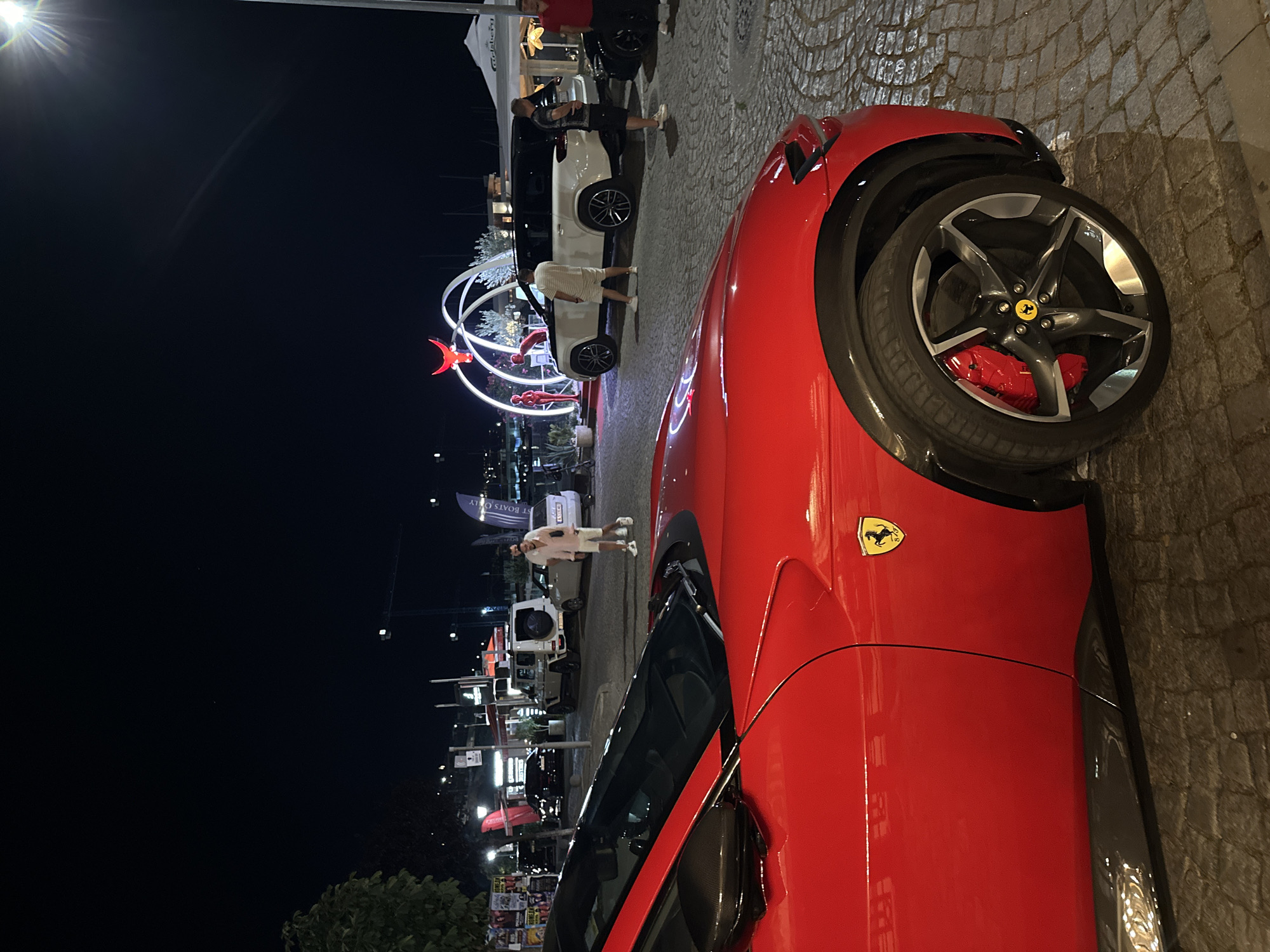 A red Ferrari is parked on a cobblestone street at night, its side and front wheel visible. In the background, there are several other vehicles, including a white SUV, a black car, and a white sedan. A few people are standing around, some near the vehicles and others in the distance. There are also some structures in the background, including what appears to be a stage or display area with a red bull sculpture and arched lights. A white tent with the "Carlsberg" logo is also visible. The location is identified as Sozopol, Bulgaria.