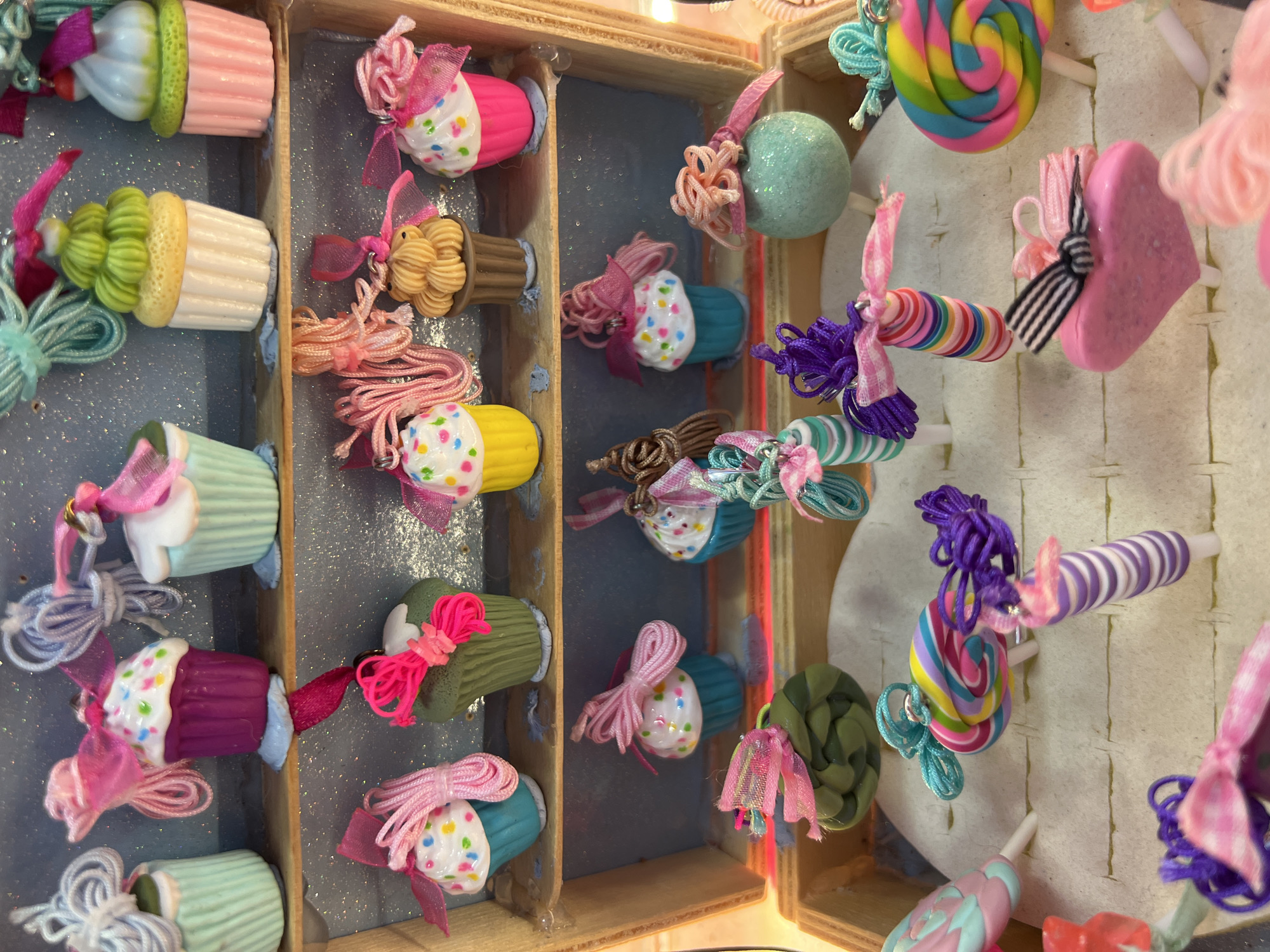 The image depicts a display case filled with novelty jewelry, primarily cupcake-themed rings and charms. The items are arranged on two wooden shelves and a base layer of ring holders. The background is a glittery blue surface. The jewelry features bright, pastel colors and is adorned with ribbons and strings of yarn, mimicking frosting and decorative elements of cupcakes and lollipops. Specific items include various cupcake designs in different colors, multicolor swirl lollipops, striped lollipop rings, and heart-shaped rings. There are no people visible in the image. The overall presentation suggests a shop or market stall selling whimsical accessories.