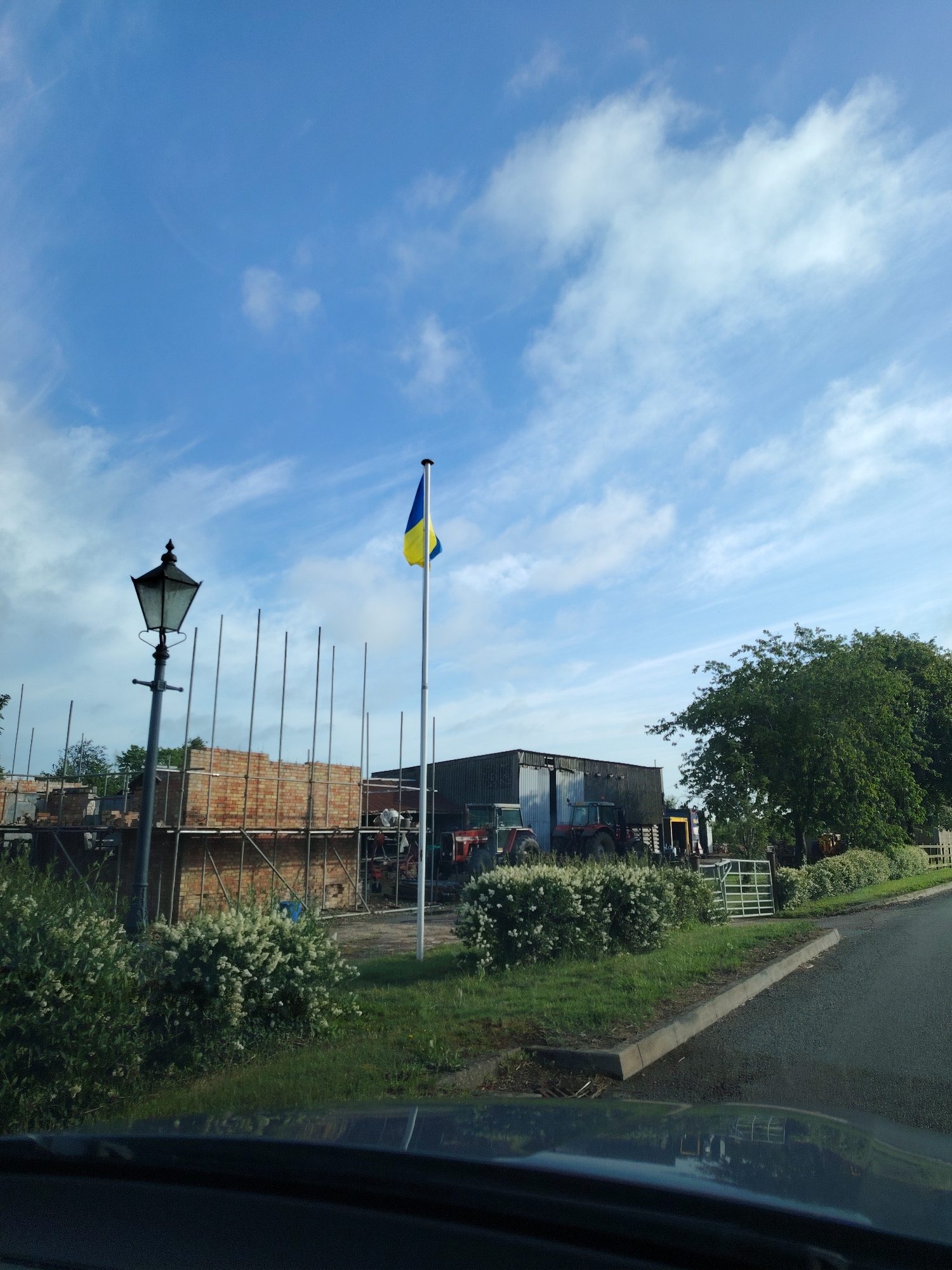 This image, captured in Huntingdonshire, United Kingdom, presents a rural or semi-rural landscape under a bright blue sky with scattered white clouds. The photograph appears to be taken from inside a vehicle, indicated by the dashboard and windshield reflection visible in the lower portion of the frame.
On the left side of the mid-ground, a tall, dark, ornate lamppost stands. Behind it, a construction area is visible, featuring scaffolding and partially constructed brick walls made of reddish-brown bricks. To the right, a prominent silver flagpole displays a Ukrainian flag, distinguished by its blue upper stripe and yellow lower stripe, partially unfurled.
Further to the right, a large barn or agricultural building with dark, corrugated metal siding is present. Parked in front of this structure are at least two red tractors. A sizable green deciduous tree with dense foliage occupies the right side of the scene. The roadside features green grass and bushes adorned with white blossoms. A concrete curb delineates the edge of an asphalt road that curves slightly to the right. A white metal farm gate is partially visible on the far right. No individuals are discernible in the image.