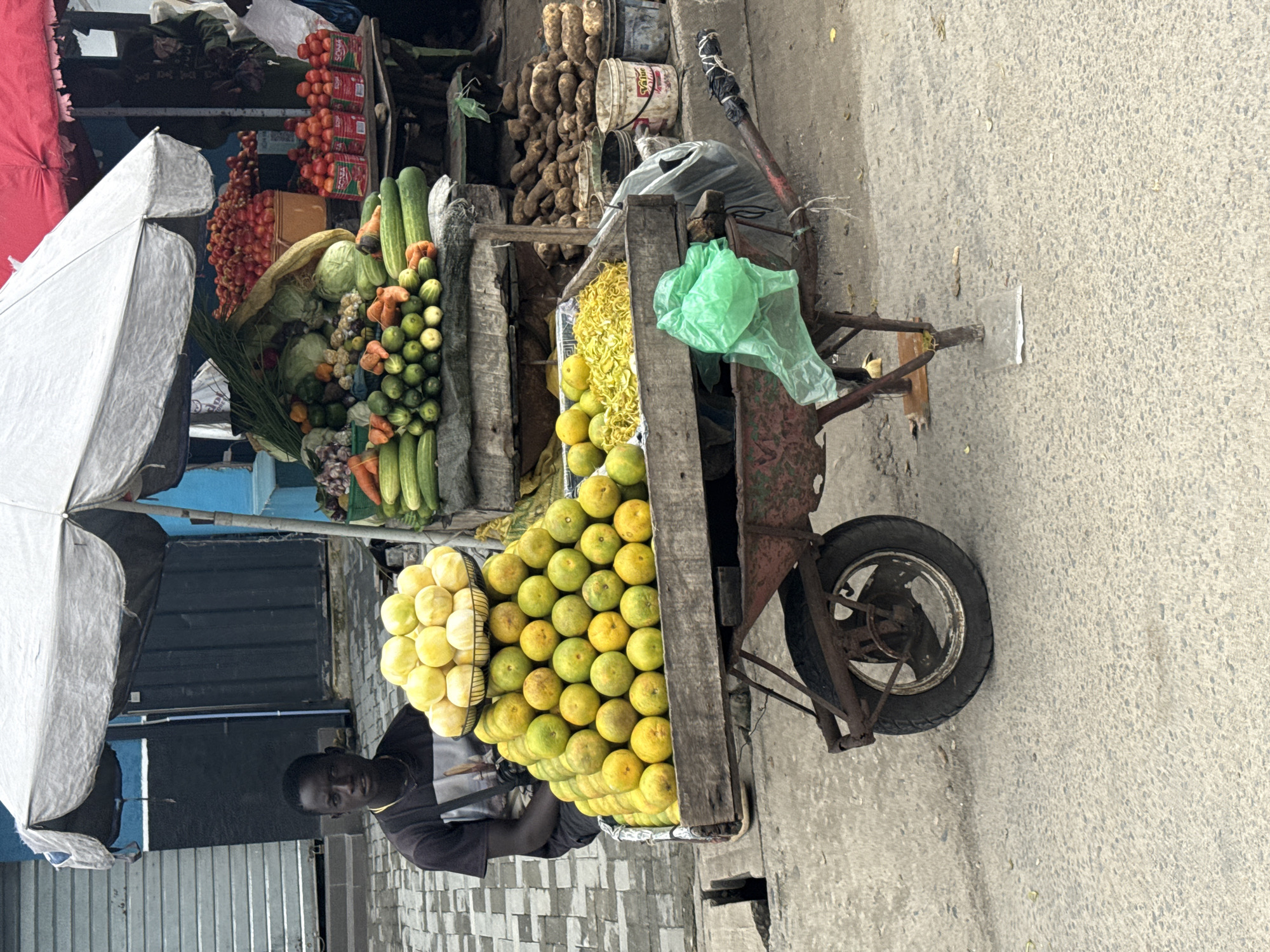 A young Black male sits behind a cart laden with fruits and vegetables at an outdoor market stall. The cart is a weathered, red wheelbarrow filled with produce. In the foreground, the wheelbarrow's wheel is visible, with its rusty spokes and worn tire. Above the wheelbarrow, large pyramids of oranges and pale yellow fruits, possibly lemons or limes, are stacked in baskets and on a wooden platform. Behind the fruit, there's a wooden crate filled with bright yellow, shredded material, possibly dried corn or plantain chips. Further back, more vegetables like cucumbers, carrots, and cabbages are displayed on shelves under a grey umbrella. In the background, red tomatoes are piled high in buckets. A dark-colored building with roll-up doors is visible behind the stall, and a blue wall is to the right. The ground is paved with concrete. The young man is wearing a black t-shirt with a graphic design. He is looking directly at the camera.