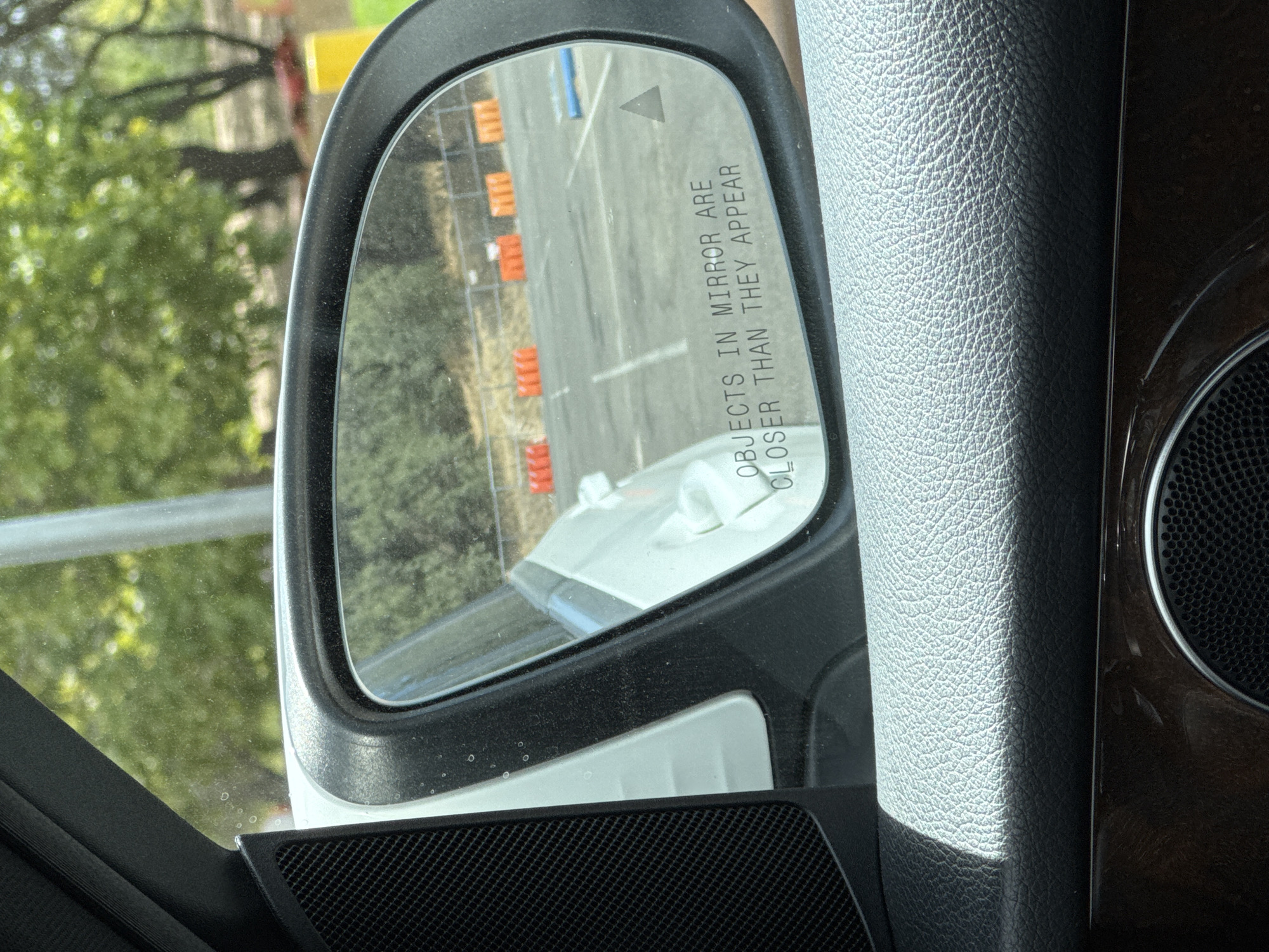 The image shows a view from inside a car, looking at the side mirror. The side mirror reflects a parking lot with orange traffic cones and white lines. The words "OBJECTS IN MIRROR ARE CLOSER THAN THEY APPEAR" are visible on the mirror. The interior of the car, including part of the dashboard and a speaker, is also visible. The exterior environment outside the car consists of trees and a pole. The image appears to be taken in Folsom, United States, given the provided context, though this specific location cannot be definitively identified from the visual information alone.