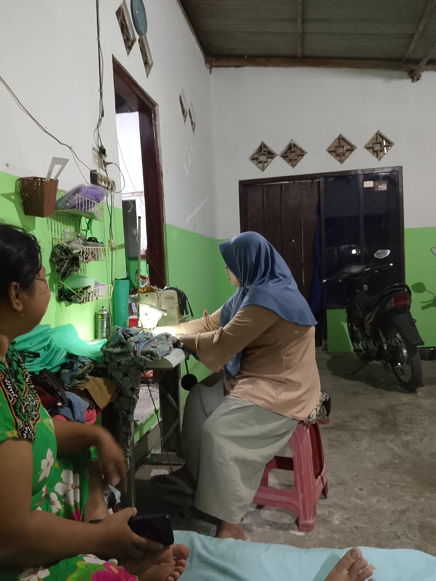 In Kelurahan, Indonesia, a woman wearing a blue hijab and a light brown shirt is sewing with a white sewing machine in a room. She is sitting on a red stool. Another woman, wearing a green floral-patterned dress, is sitting on the left side of the frame, looking at her phone. Various fabrics and sewing supplies are scattered around the sewing machine and on shelves on the wall. A motorcycle is parked outside a doorway in the background, suggesting it is a residential or small workshop setting. The walls are painted white and light green, and the floor is concrete. The scene appears to be illuminated by artificial lighting, as it is dark outside.