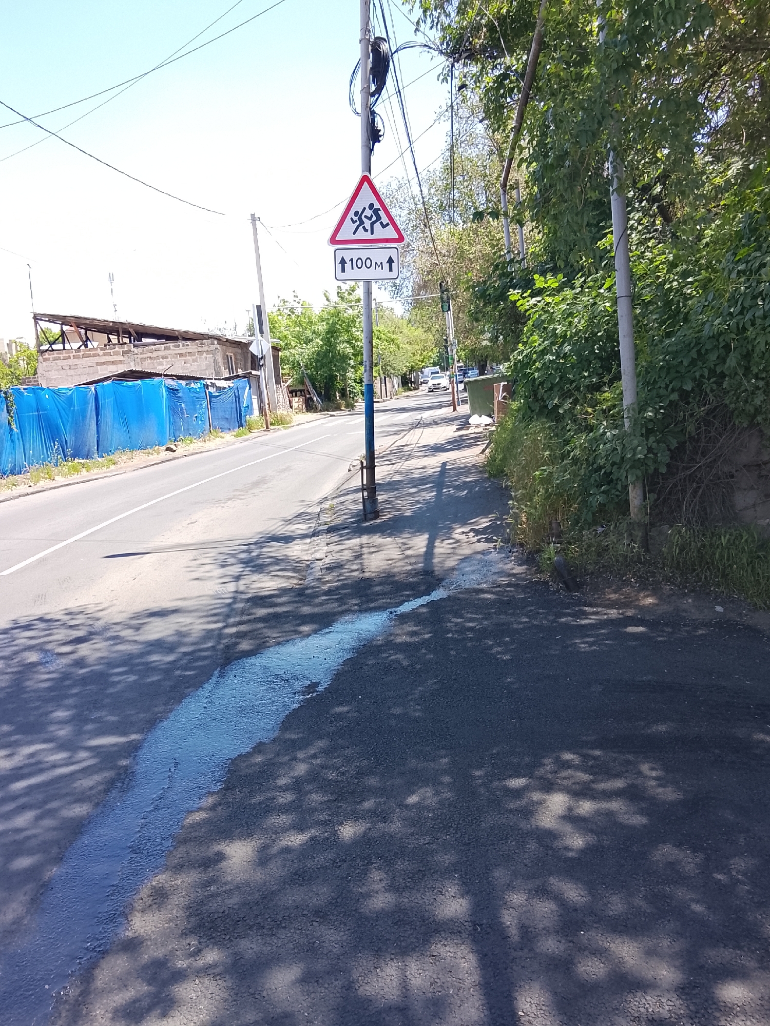The image captures a sunlit outdoor scene of a road and an adjacent sidewalk, likely in a semi-urban or residential area. The perspective is from the sidewalk, looking forward along the road.
On the right side of the image, a paved sidewalk is bordered by dense green foliage and trees, some of which are growing against a stone or concrete wall. Several utility poles with electrical wires run parallel to the road. Prominently featured is a traffic sign mounted on a pole: a red-bordered white triangle depicting two running human figures, indicating a "children crossing" or "children ahead" warning. Below it, a rectangular white sign has two upward-pointing arrows flanking the text "100M", signifying that the warned area is 100 meters ahead.
The asphalt road to the left of the sidewalk has a white dividing line running down its center. On the left side of the road, a long section of blue tarp or fencing conceals an area, behind which a partially constructed or dilapidated building with an exposed roof structure is visible. Further down the road in the distance, a few parked white cars and a green dumpster can be seen.
The foreground of the image features strong, dappled shadows from overhead trees cast across the road and sidewalk. A distinct light blue, glossy streak, possibly spilled paint or some other liquid, runs along the edge of the asphalt in the bottom-left corner of the frame. The sky is bright and clear, indicating a sunny day, likely midday or early afternoon given the angle and intensity of the shadows. No people or active events are visible in the static scene.