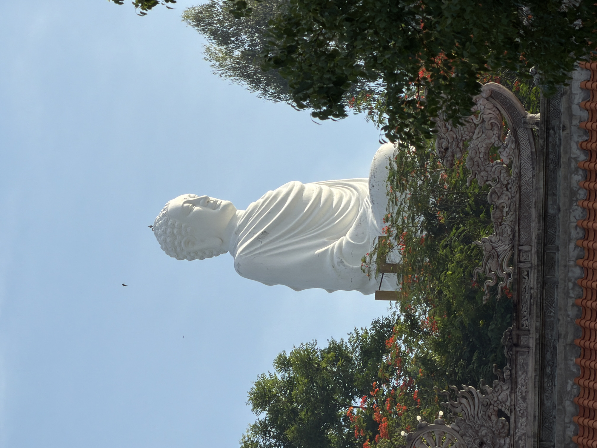 The image shows a large, white, seated Buddha statue, viewed from a low angle, with a clear blue sky as the background. The statue is partially obscured by lush green trees and flowering plants with bright orange blossoms. In the foreground, there is ornate architectural detailing, including what appears to be a tiled roof and decorative stone carvings of dragons and a Dharma wheel. The statue is located in Nha Trang, Vietnam, at the Long Son Pagoda. The scene is peaceful and serene, bathed in natural light. No people are visible in the image.