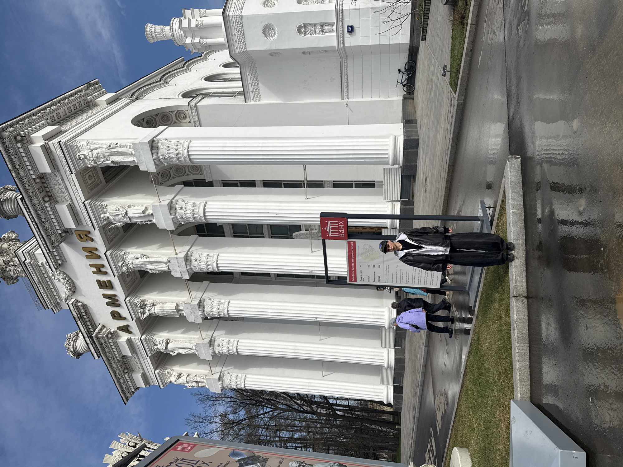 The image shows the facade of the "Armenia" pavilion in the VDNH exhibition park in Moscow, Russia. The pavilion is a white building with classical architectural elements, including columns and sculptures. The word "ARMENIA" is visible above the entrance. Several people are standing in front of the building. A man in a black jacket and cap is standing near a sign. Two other people are walking past. The ground appears wet, reflecting the surroundings. A bicycle is parked near the building.