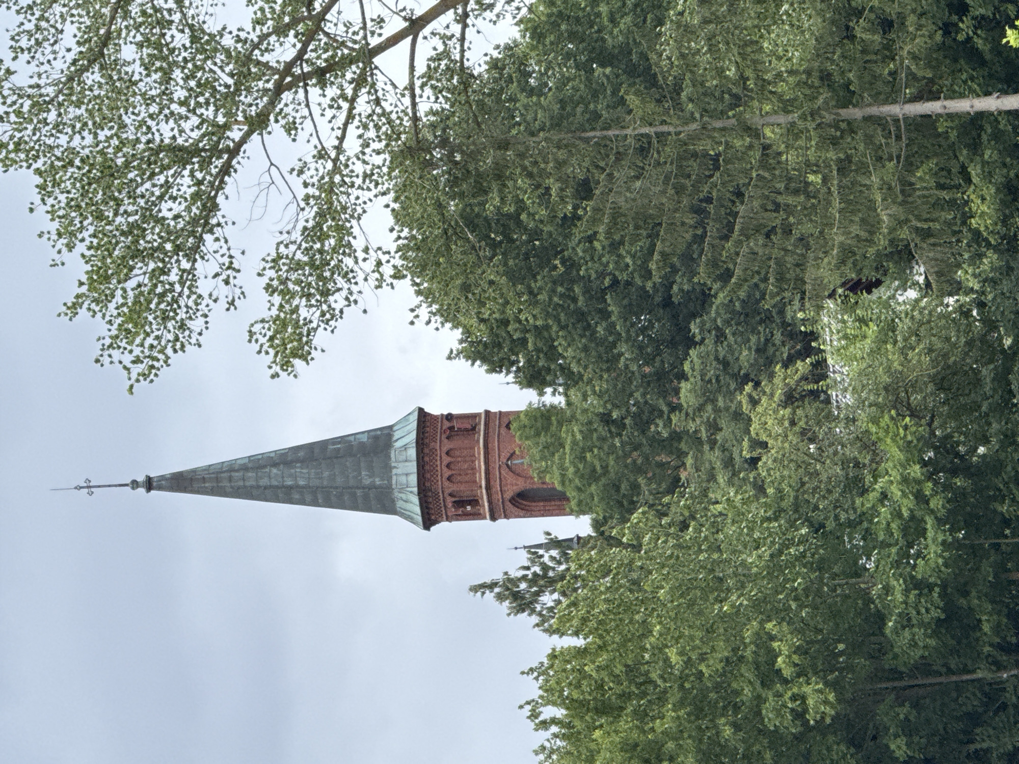 The image shows a church steeple emerging from a dense canopy of green trees. The steeple is constructed of red brick with a dark, possibly copper or slate, roof that tapers to a point topped with a finial and cross. The steeple has arched windows and decorative brickwork near its peak. Several trees of varying types and sizes dominate the foreground and surround the church, obscuring much of the building. The sky is overcast and a pale grey. The scene is located in Kołczewo, Poland. There are no people visible in the image.