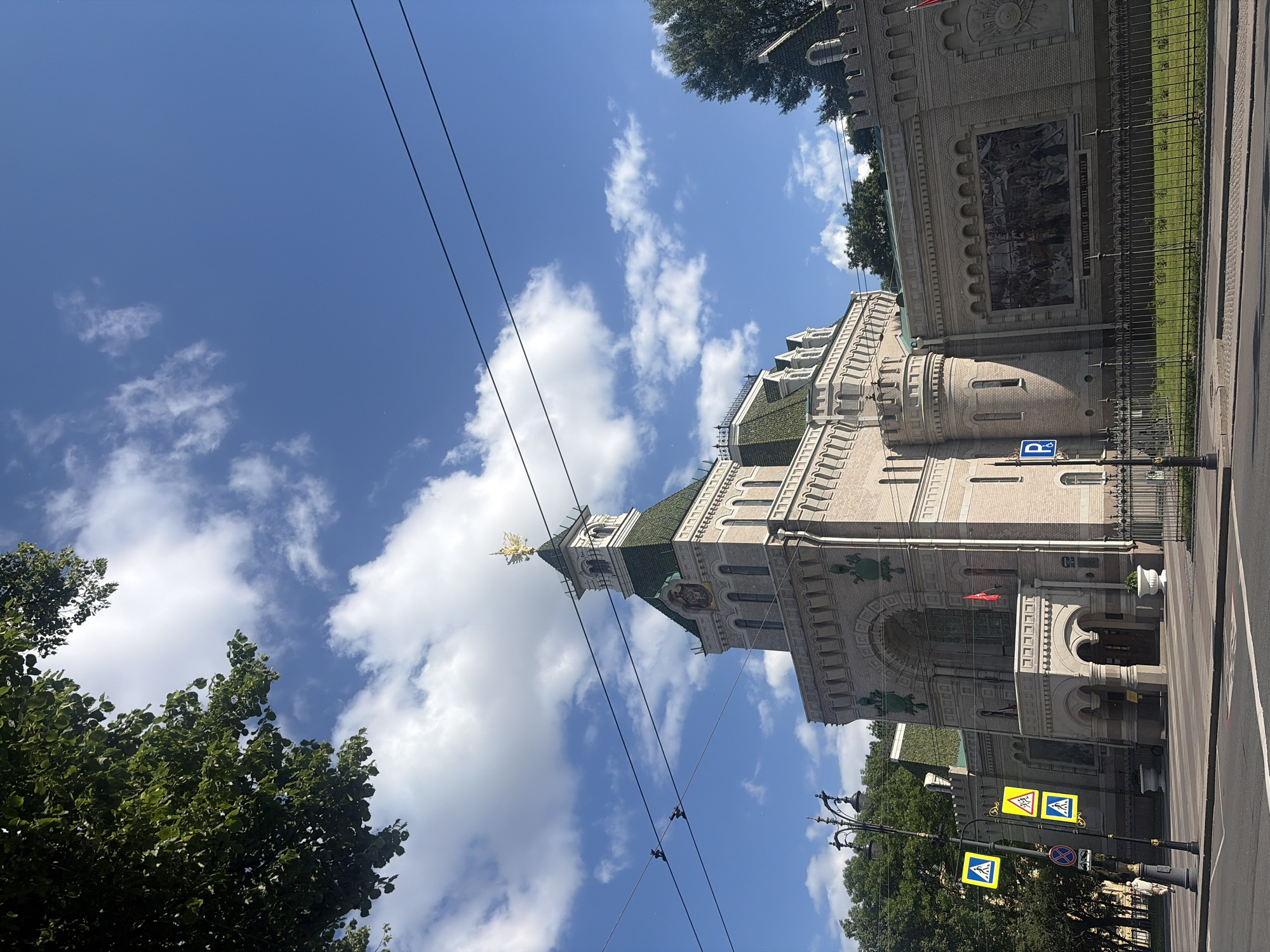 The image shows a wide shot of the historic building, the Kazan Barracks, located in Saint Petersburg, Russia. The building features a light-colored brick facade with arched windows and decorative elements. A prominent tower with a green roof and a golden double-headed eagle emblem stands out at the top. The sky is bright blue with scattered white clouds. In front of the building, a street with pedestrian crossings and traffic signals is visible. The scene is captured from across the street, with a portion of a lush green tree in the upper left corner and more trees to the right. There are power lines strung across the sky. A mural depicting a historical scene is mounted on a wall of the building. A black metal fence separates the building from the sidewalk and street.