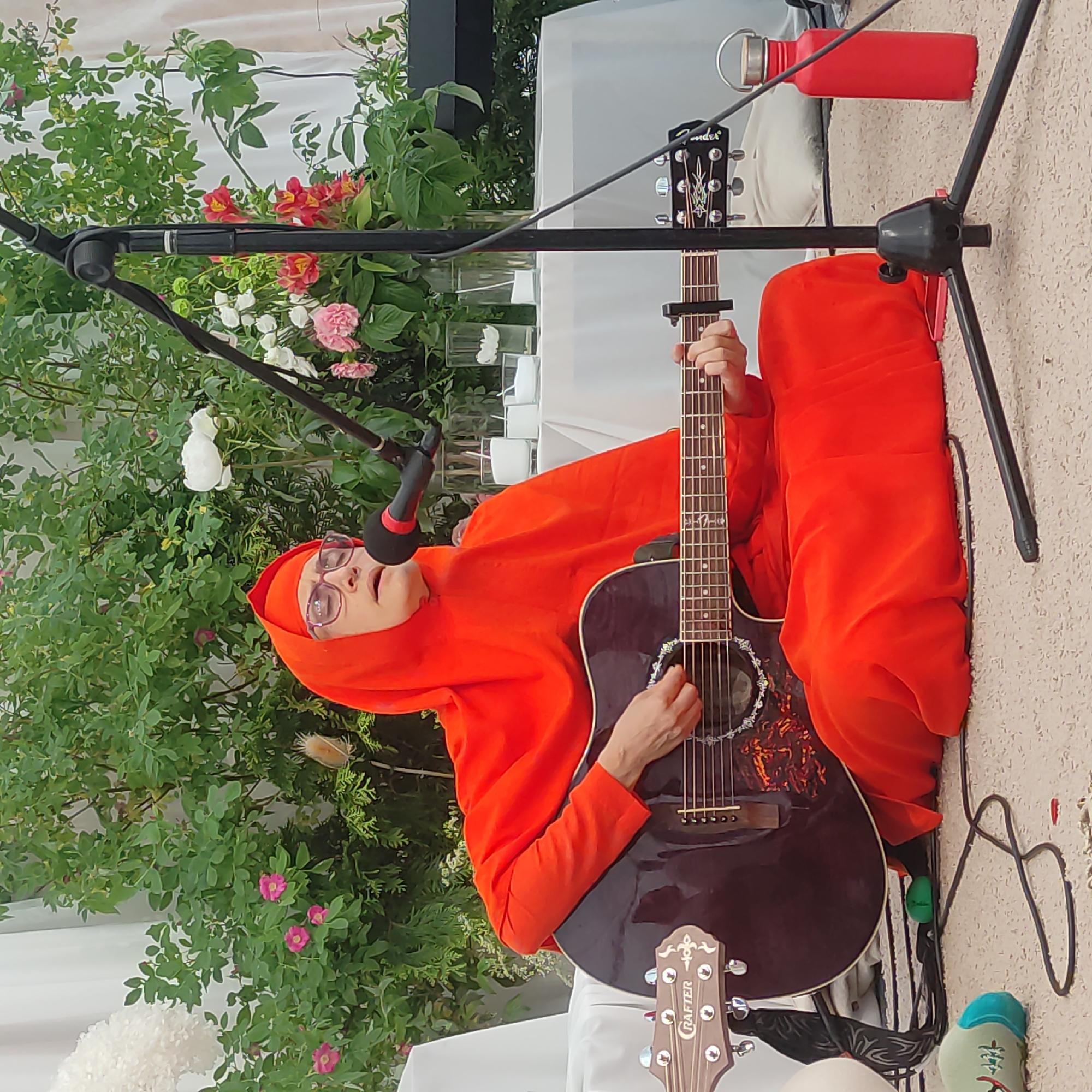 A person wearing an orange hooded robe and glasses is seated on a light-colored carpet, playing an acoustic guitar and singing into a microphone. The guitar is dark brown with a light-colored pickguard. Behind the person is a table covered with a white cloth, on which are several clear glass vases containing white candles. Behind the table, a dense green bush with pink and white flowers is visible. To the right of the scene, a black microphone stand is positioned, and next to it, a red water bottle stands upright. To the left of the frame, the headstock of another guitar is visible, labeled "RAafter". The environment appears to be outdoors, possibly in a garden or park setting. The image was captured in Zhirnosovo, Russia.