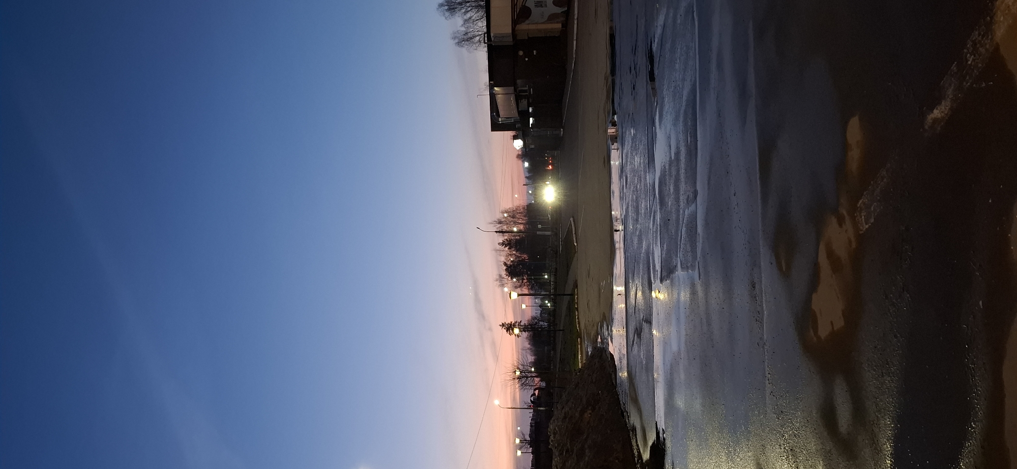 The image captures a street scene in Chaikovsky, Russia, at twilight. The sky transitions from deep blue to orange near the horizon. Along the street, numerous illuminated lampposts are visible, along with trees, a building, and a wet, reflective road surface. The scene is absent of people, focusing instead on the infrastructure and the atmospheric conditions. The lighting suggests early evening, with the street lights compensating for the diminishing natural light. The wet road creates reflections, doubling the visibility of the street lights and adding depth to the image.