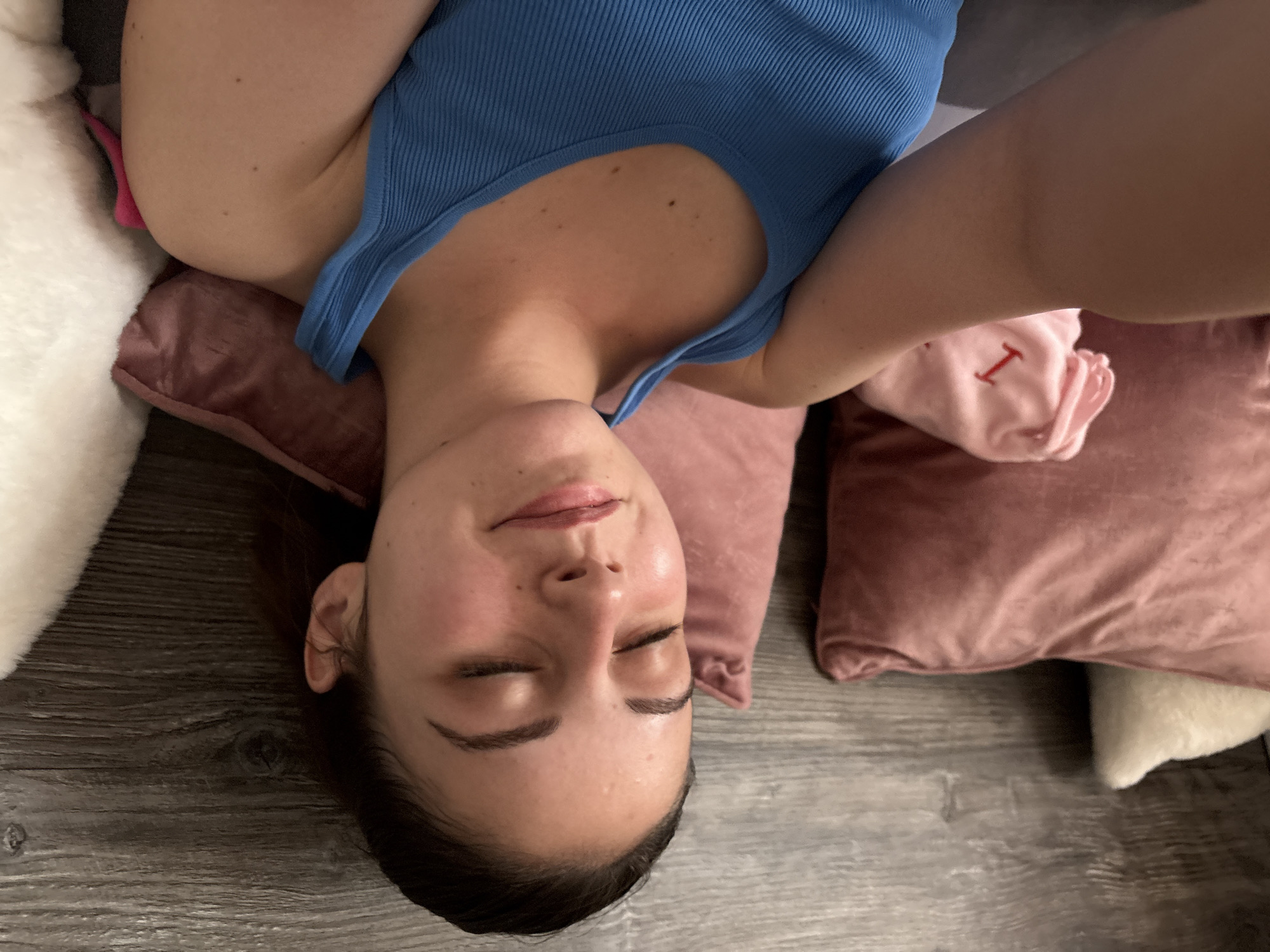 A woman is lying down with her eyes closed, appearing relaxed. She is wearing a blue tank top. Her head rests on a pink pillow, and there is a white fluffy object and another pink pillow nearby. The flooring beneath her appears to be gray wood-like planks. The background is blurred, obscuring any details about the location; the image does not provide any information about the city of Bulgarovo, Bulgaria.