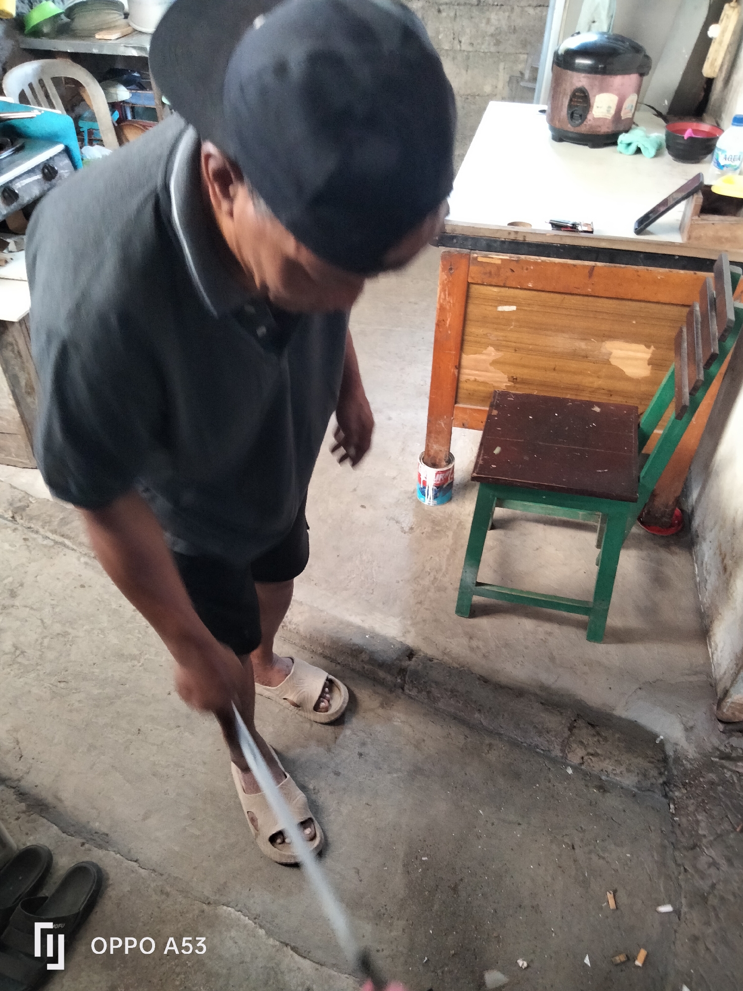 In Denpasar, Indonesia, a man wearing a dark t-shirt, black shorts, a black baseball cap, and light-colored sandals is seen sweeping the concrete floor with a broom. The floor appears to be outdoors, possibly in a yard or alleyway, as indicated by the rough concrete surface and the presence of several cigarette butts scattered on the ground. To the right of the man, a small green chair with a dark wooden seat is positioned near a wooden structure. In the background, a rice cooker and other items are visible on a table. The scene is dimly lit, suggesting it might be indoors or an area with limited natural light. The overall impression is one of a mundane, everyday activity taking place in a functional, somewhat cluttered environment.