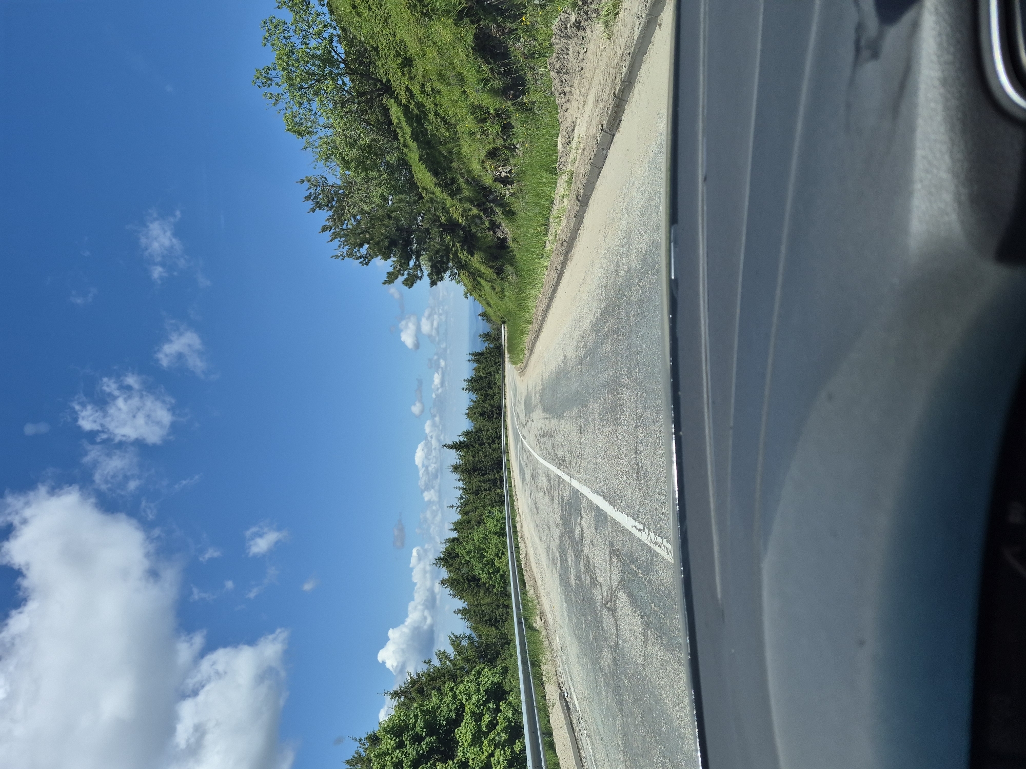 The image captures a scenic view from inside a vehicle, looking out at a winding mountain road in Beli Osam, Bulgaria. The road, paved and marked with a white line, curves to the right, flanked by a metal guardrail on the left and a lush green hillside on the right. Dense evergreen trees form a forest on the left side of the road, while the hillside to the right is covered in green grass and scattered trees. The sky above is a vibrant blue, dotted with fluffy white clouds. The foreground of the image shows the dashboard of the vehicle, indicating that the photo was taken while driving. The overall impression is one of a serene and picturesque drive through a natural landscape.