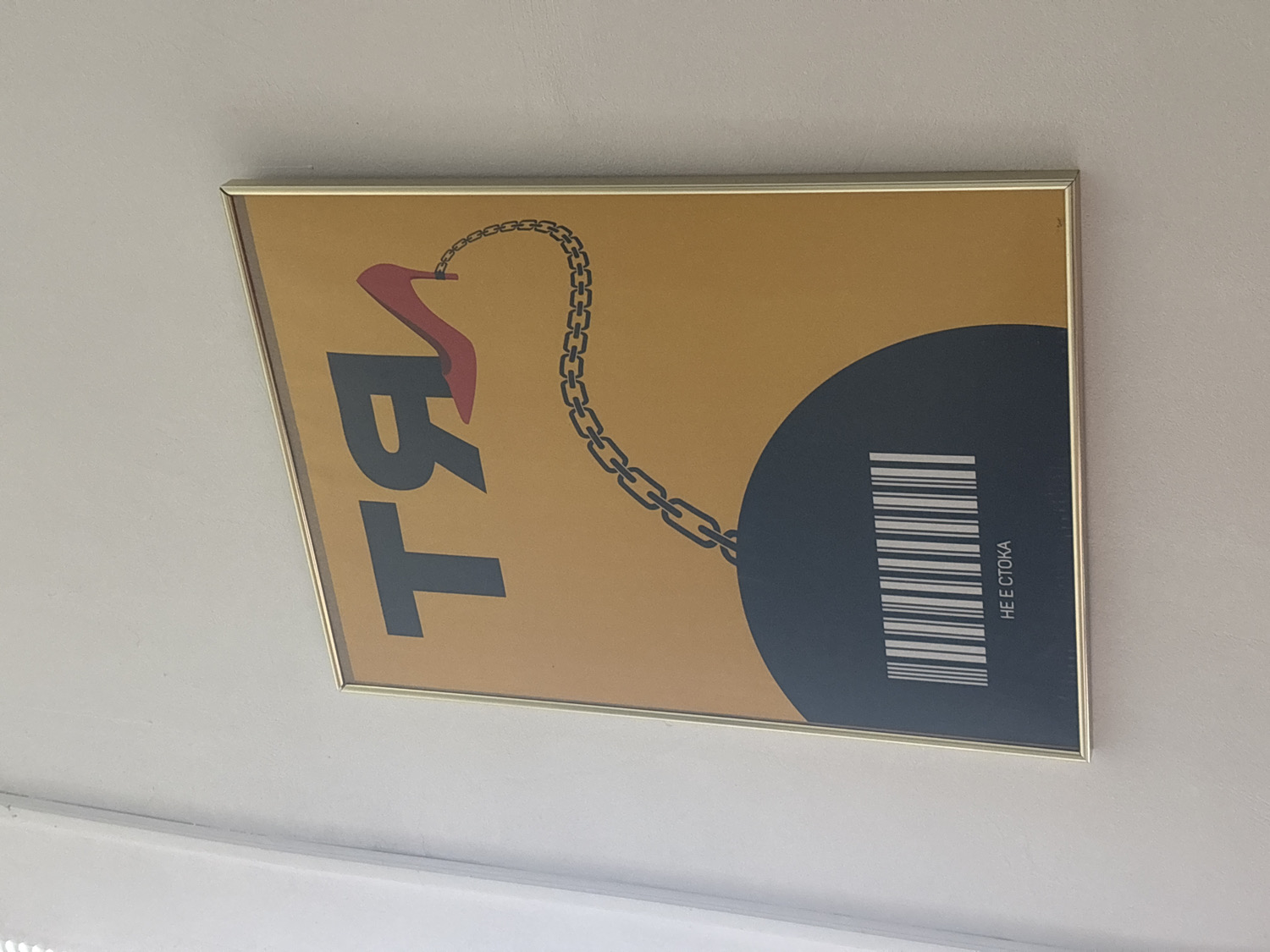 A framed poster is captured from a slightly angled perspective, hanging on a plain, light-colored wall. The artwork, which appears to be modern or graphic design, features a warm yellow background. Prominently displayed on the left are large, stylized letters in dark gray, forming "TЯ" (Cyrillic for "She"). A vibrant red high-heeled shoe is depicted near the top of the "Я", connected by a dark chain that curves across the poster.
The chain extends to a large, dark blue or black circular shape located in the bottom right section of the poster. Within this circular shape, a barcode pattern is visible in white. Oriented vertically alongside the barcode are the Cyrillic words "НЕ Е СТОКА". This phrase translates to "Is not merchandise" or "Is not a commodity". The overall composition, with the high heel chained to a barcode-laden sphere and the explicit text, suggests a powerful social commentary on women's objectification or commodification. The frame around the poster is thin and metallic, possibly gold or brass.