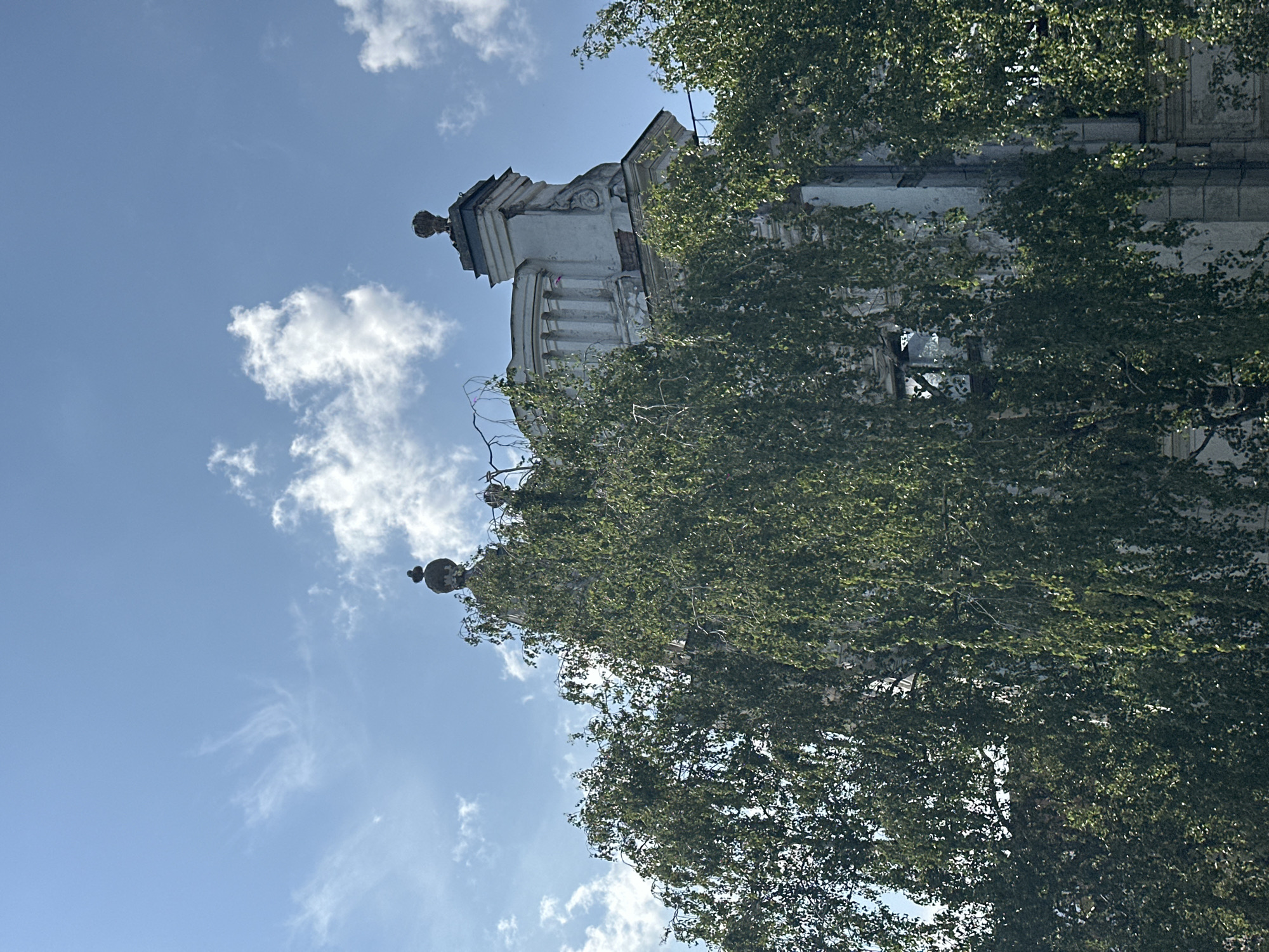 This is a low-angle, eye-level shot of an old, weathered building facade peeking through lush green trees under a bright blue sky with wispy white clouds. The building appears to be made of stone and is partially obscured by the foliage of several large trees, possibly birch trees, due to their thin, light-colored bark and delicate leaves. The building's architecture suggests it is from a historical period, with visible decorative elements like columns and a domed structure on top, topped with a finial. One section of the building shows signs of decay, with exposed brickwork and what appears to be a broken window. The scene is set in the city of Biysk, Russia, likely on a sunny day. There are no people visible in the image.