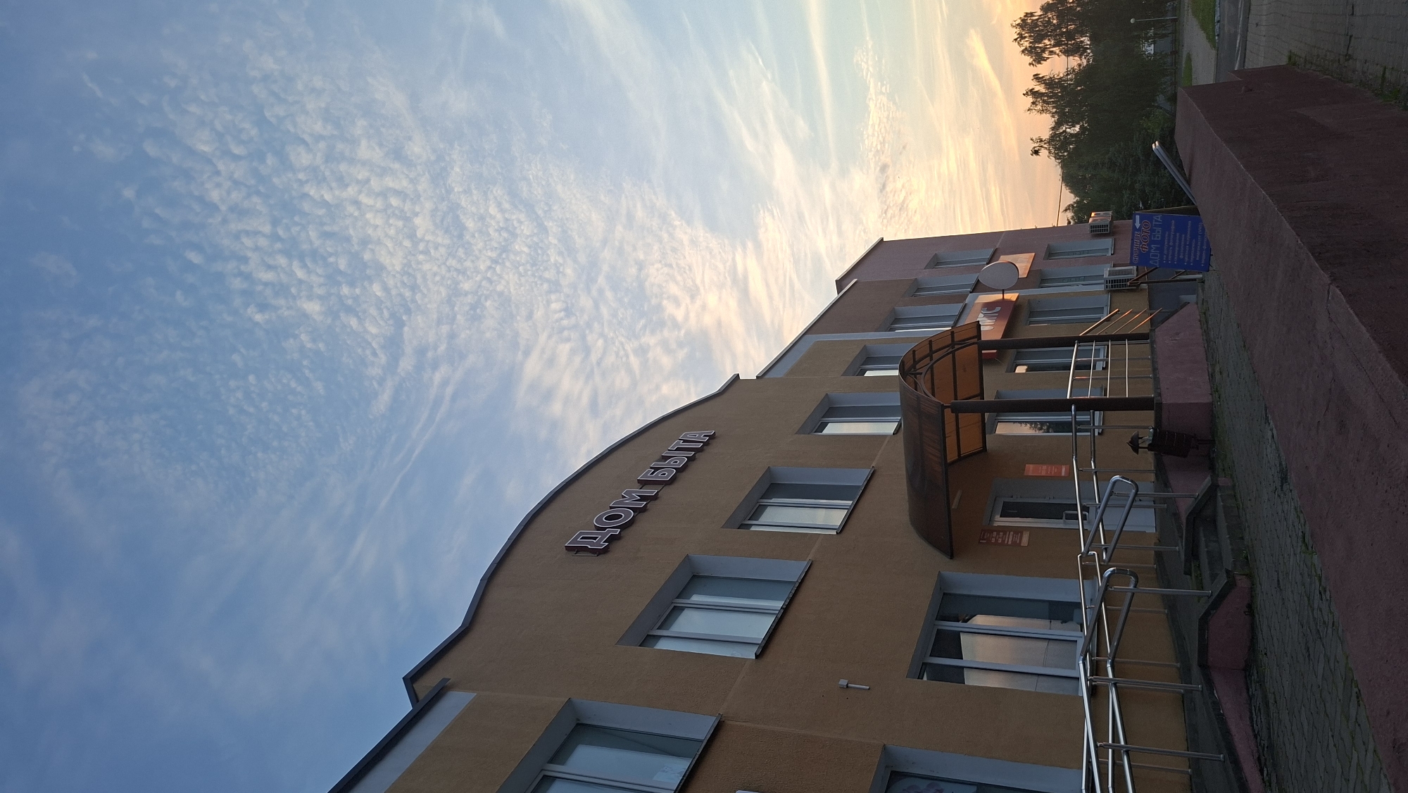 The image shows a multi-story building with an orange facade and a sign that reads "ДОМ БЫТА" (House of Services) in large, illuminated letters. The building has several windows and an awning over the entrance. The sky above is a clear blue with scattered clouds, suggesting it is daytime. In the foreground, there is a paved walkway and a low, dark red wall. Trees are visible in the background, as well as another building with a pinkish hue. A blue sign with Cyrillic text is posted near the entrance, which appears to advertise services. The overall scene depicts a commercial building in what seems to be a suburban or urban setting in Myadzyel, Belarus. There are no people visible in the image.