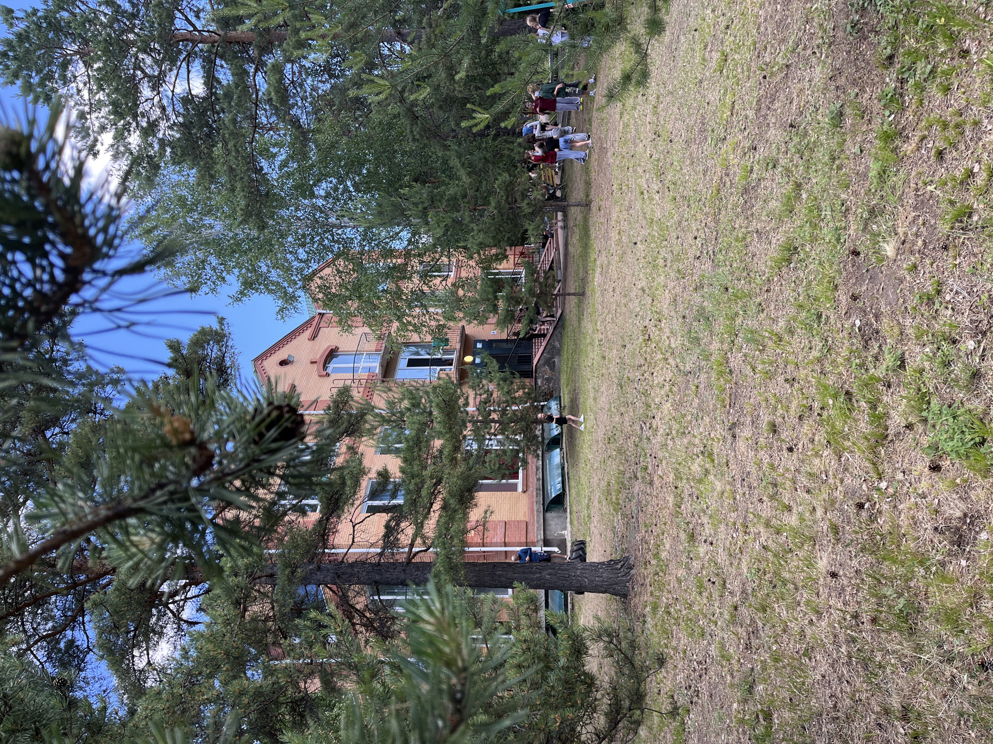 A three-story brick building with a red tile roof stands amidst a grove of pine and birch trees. The building has several windows and balconies, and a red-brick chimney. In front of the building, a group of young people are gathered on a grassy lawn dotted with pine cones and scattered needles. Some individuals are standing and talking, while others appear to be walking or playing. The sky above is blue with scattered white clouds. The overall scene suggests an outdoor recreational area or a summer camp in the city of Krasnoturansk, Russia.