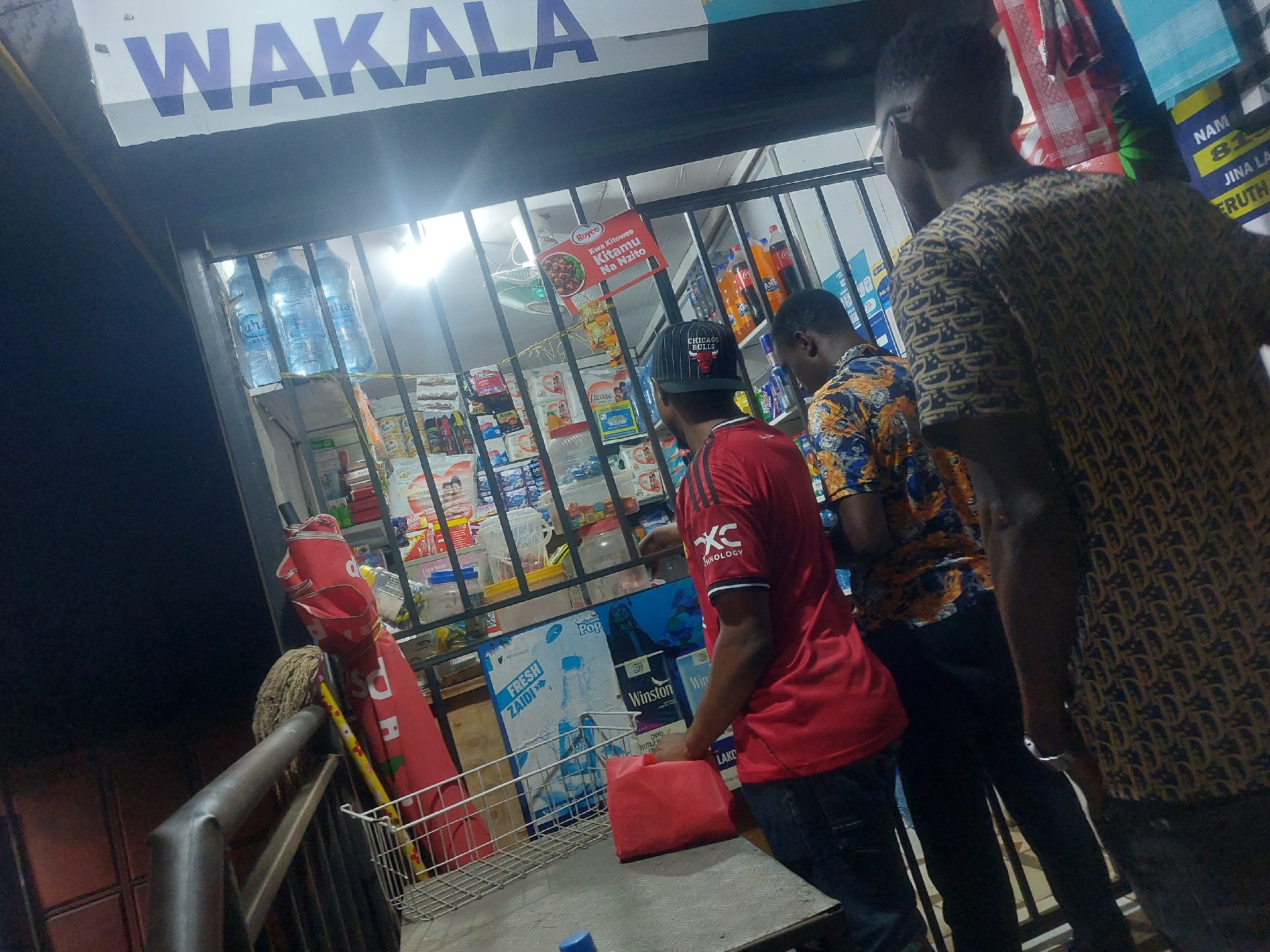 The image shows three men standing in front of a small shop or kiosk, likely a local "duka," in what appears to be a residential area in Dar es-Salaam, Tanzania. The shop, labeled "WAKALA," sells a variety of goods including bottled water, beverages, and other household items. The shop is enclosed by metal bars. The men are dressed in casual clothing. One wears a red shirt and a black Chicago Bulls hat and carries a red plastic bag. Another wears a patterned orange and blue shirt. The third man is partially visible, wearing a tan patterned shirt. A promotional umbrella with a red cover leans against the shop. The scene is illuminated by a bright light inside the shop, suggesting it is evening or night. Signage for products like Royco, Fresh Zaidi water, and Winston cigarettes is visible. A sign reading "NAM 815 JINA LA ERUTH" is also present.