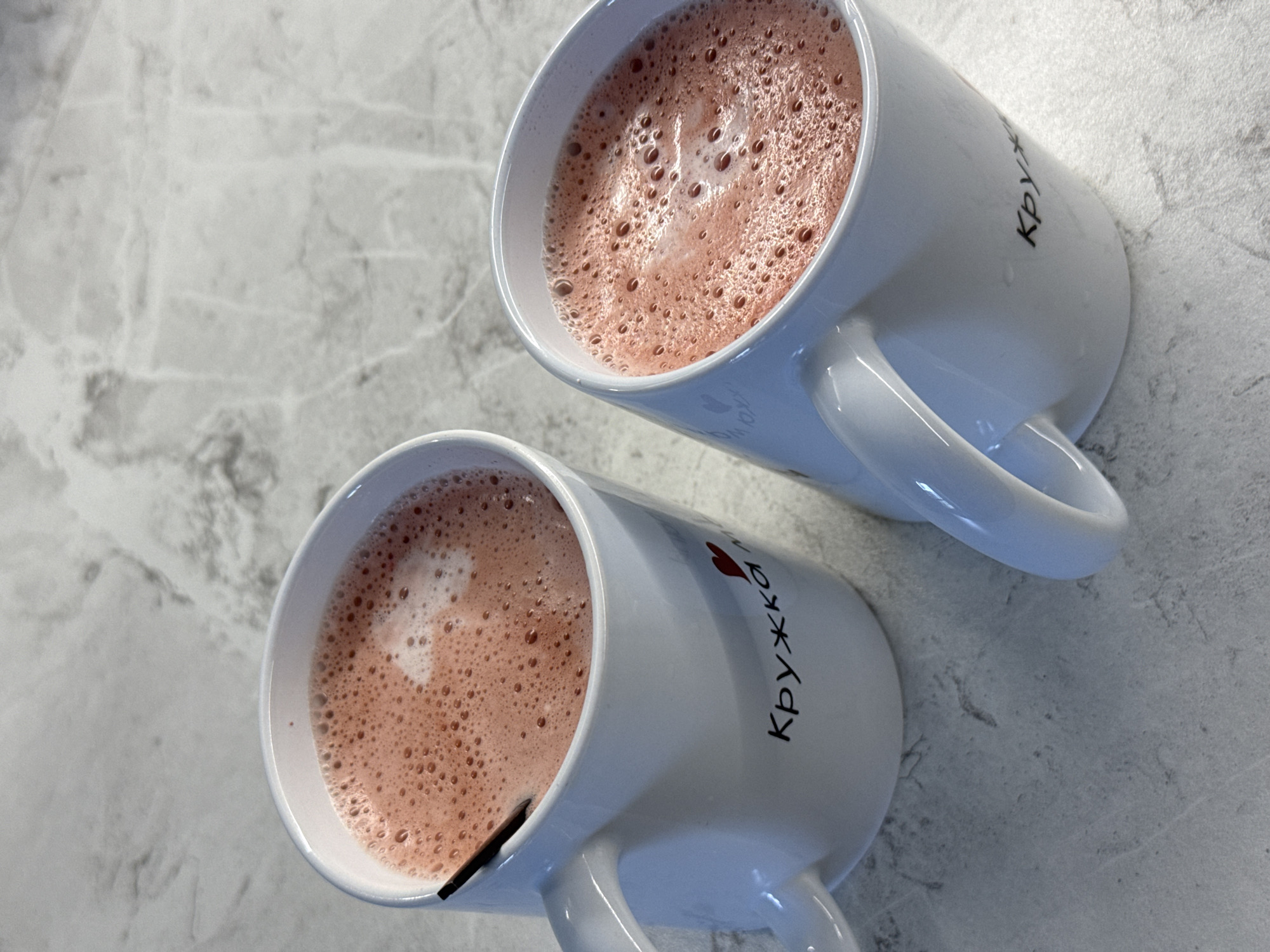 Two white ceramic mugs filled with a pinkish-red beverage topped with foam sit on a light gray speckled surface, possibly a countertop or table. A black stirring stick is visible in the mug on the left. The mug on the left has black lettering that appears to be "Кружка" and a small red heart graphic. The mug on the right also has similar black lettering, partially obscured. The surface has a marble-like pattern. There are no people visible in the image. The scene is indoors. The image is taken from a top-down perspective. There is no information about the location being in Lyubertsy, Russia, within the image itself.