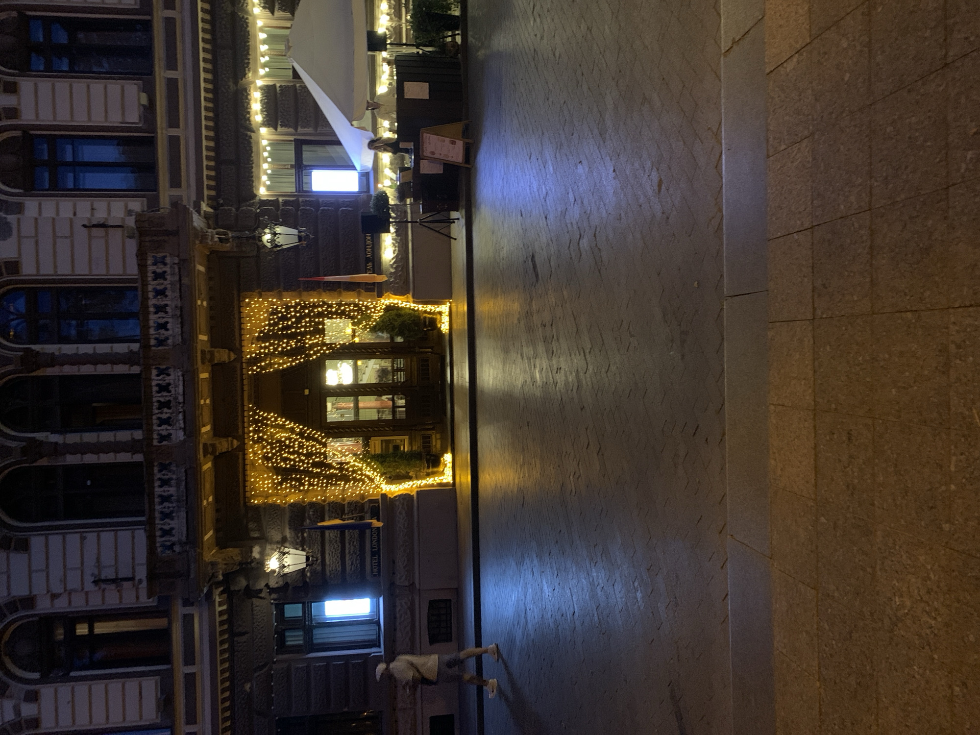 The image shows a street scene at night in Odesa, Ukraine, in front of the Hotel London. The hotel facade is visible, with ornate architectural details and arched windows. A doorway is decorated with a cascade of warm-toned string lights. To the right of the entrance, a white umbrella covers an outdoor seating area, where two individuals are present behind a counter. A person wearing a white cap and shorts is walking on the cobblestone street in the foreground. The street is illuminated by the lights of the hotel and the surrounding area, casting reflections on the wet pavement. A flag, likely the Ukrainian flag, is visible near the entrance.