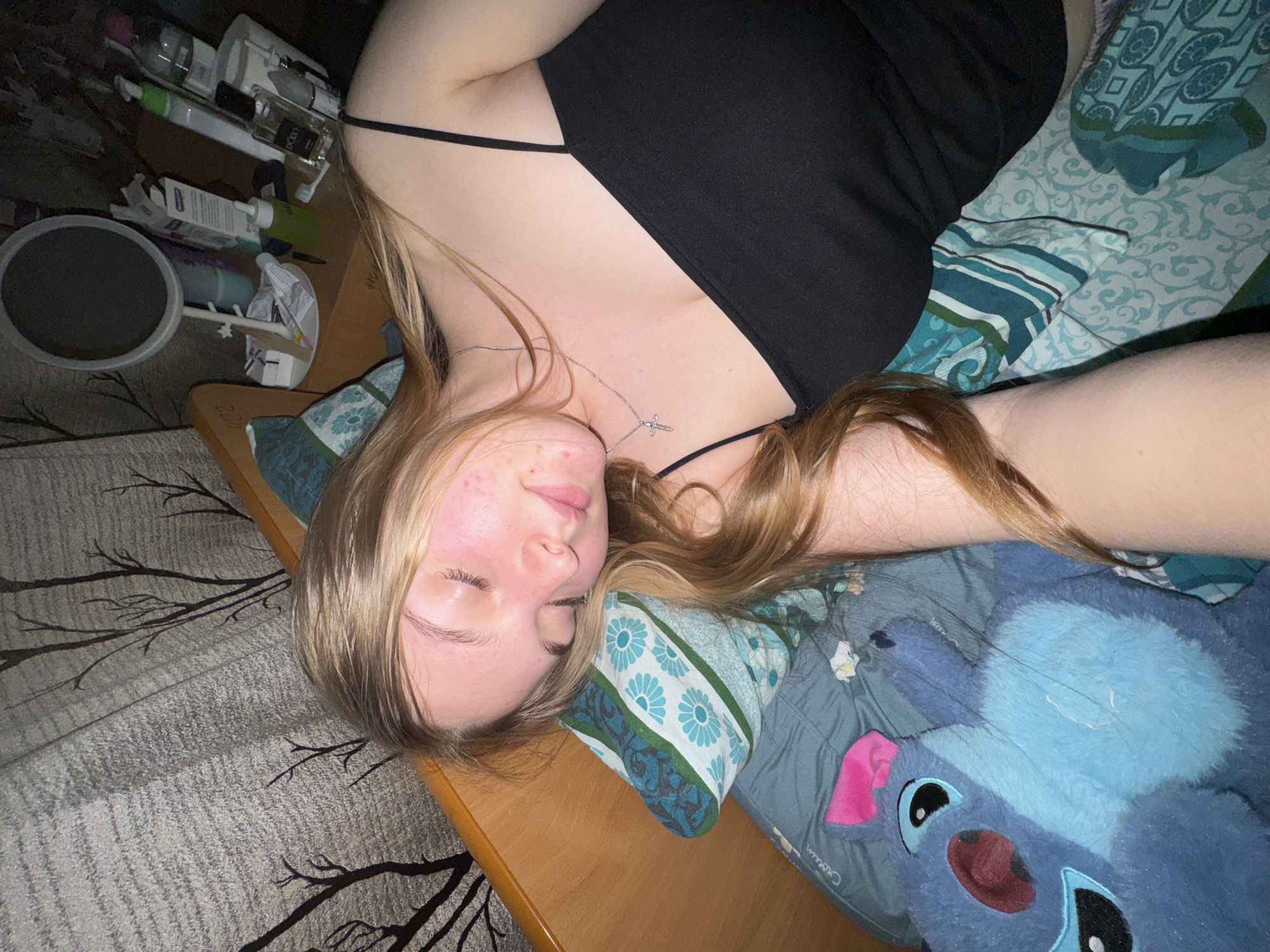 A young fair-skinned woman with long blonde hair is reclining on a bed or surface covered in blue and green patterned bedding, with a blue plush animal toy visible near her. She is wearing a black sleeveless top and a silver necklace with a cross pendant. Her eyes are closed, and her face is slightly turned to the side, displaying some blemishes on her cheek. To the left, there is a nightstand cluttered with toiletries and other personal items. The floor is covered with a patterned rug. The scene does not provide any information about the city of Perm, Russia.