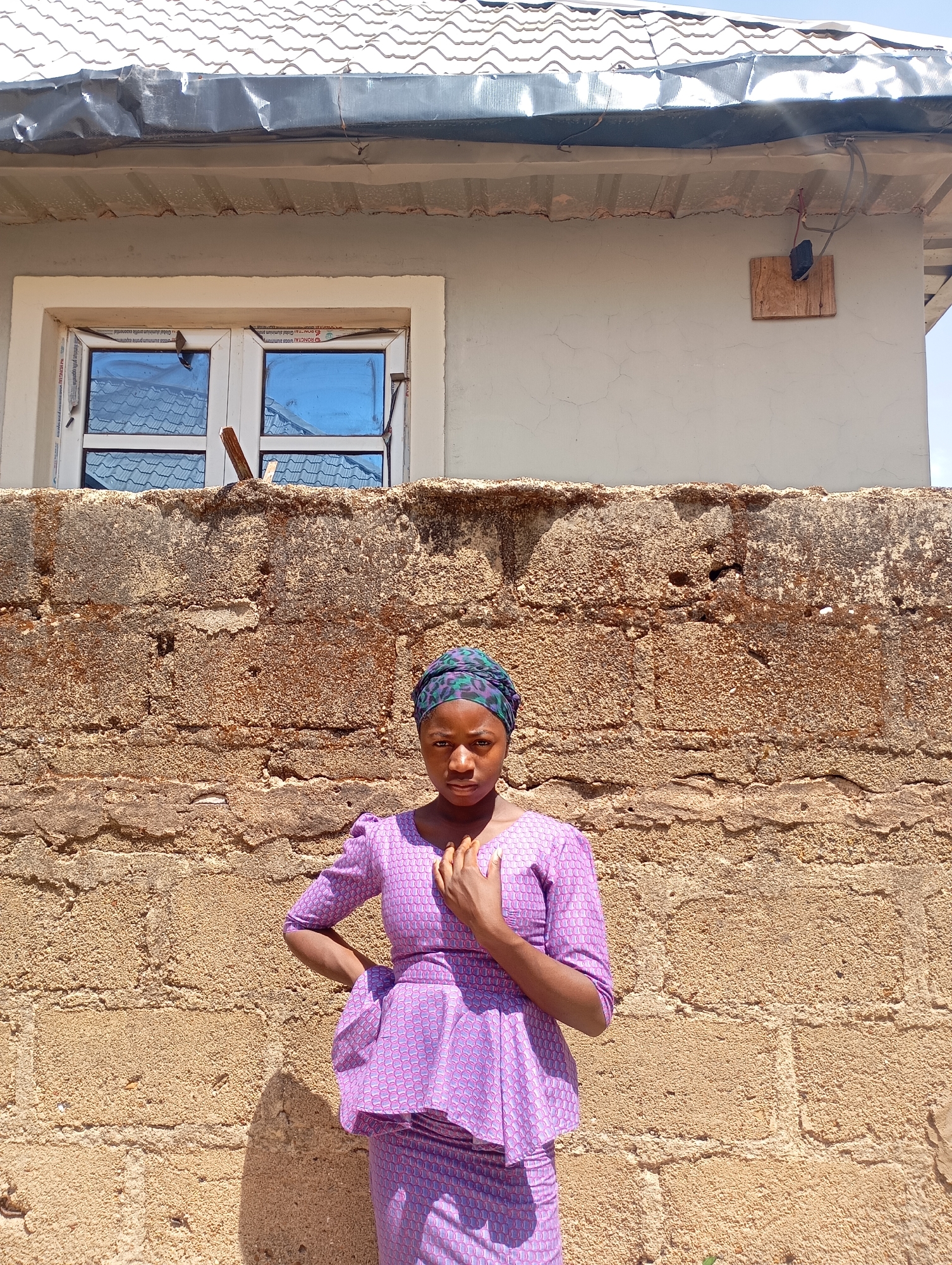 A young woman stands in front of a rough, aged brick wall, likely composed of locally sourced materials, commonly seen in older residential areas of Jos, Nigeria. She is dressed in a patterned, purple two-piece outfit, consisting of a top with short sleeves and a peplum design, and a matching skirt. A patterned headscarf adorns her head. The woman is positioned slightly off-center, with her left hand resting on her hip and her right hand placed near her chest. Behind the wall, a single-story building is visible, featuring a white-framed window with reflective blue glass and a corrugated metal roof. An electrical component is mounted on a wooden plank attached to the wall of the building. The lighting suggests it is a sunny day, casting shadows on the ground in front of the wall. The environment appears to be a residential or semi-urban area in Jos, Nigeria, indicated by the architectural style and building materials used.