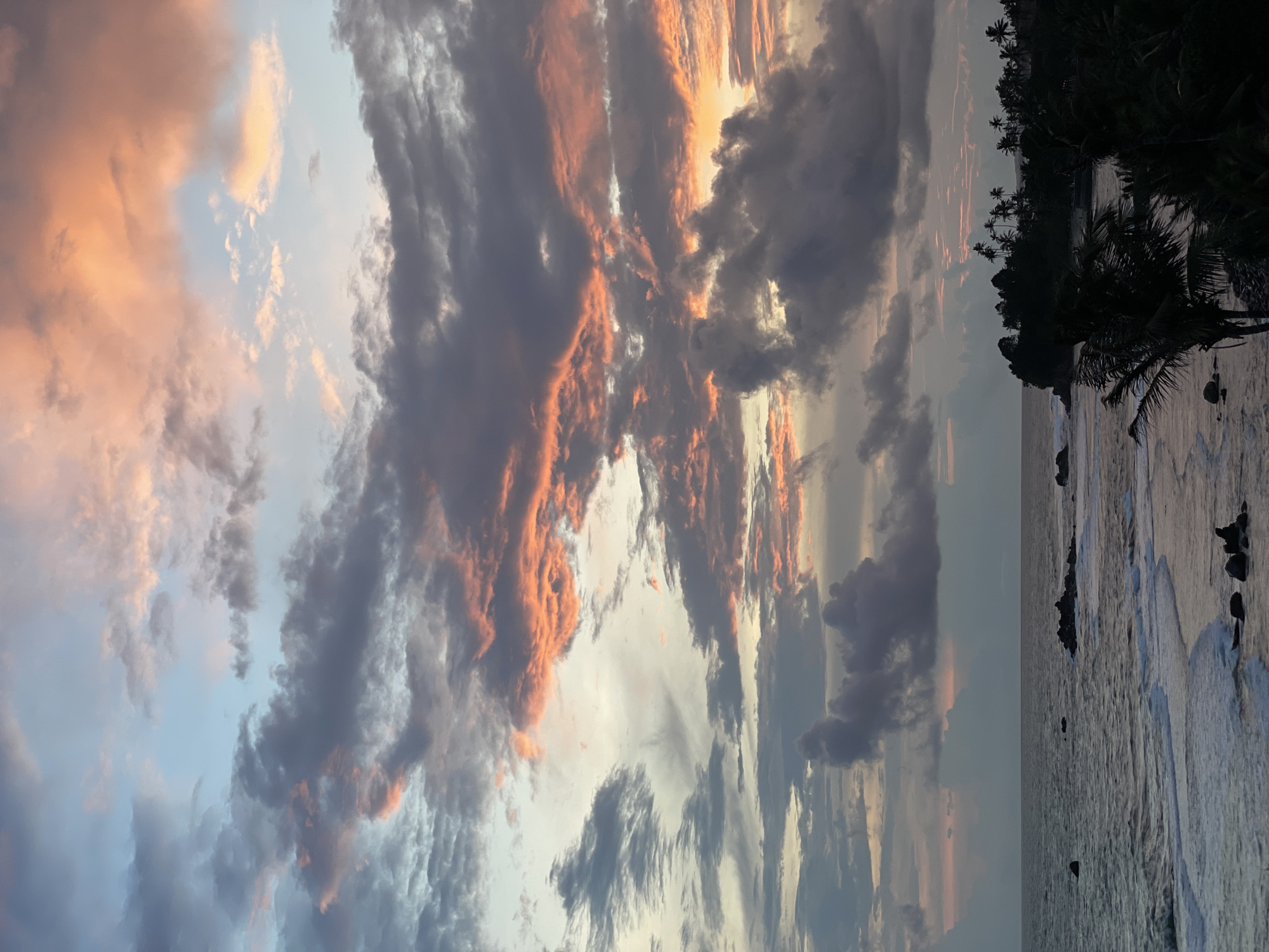 The image shows a sunset over the ocean in Goviyapana, Sri Lanka. The sky is filled with clouds, some dark gray and others tinged with orange and pink from the setting sun. The ocean is calm with gentle waves. The shoreline is visible with dark silhouettes of trees and vegetation, including palm trees. Several rocks are visible along the shoreline.