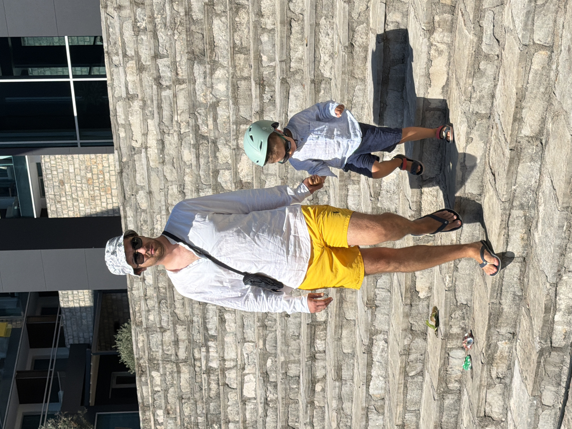 In Budva, Montenegro, a man and a young child are walking down a wide, stone staircase outdoors on a sunny day. The man, wearing a white long-sleeved shirt, yellow shorts, sunglasses, and a bucket hat, is holding the child's hand. The child, dressed in a light blue long-sleeved shirt, dark blue shorts, a light green helmet, and sunglasses, walks alongside him. The staircase is constructed from large, rough-cut stones, and there are some small toys and a green leaf scattered on the steps. In the background, a modern building with large windows and brick accents is visible.