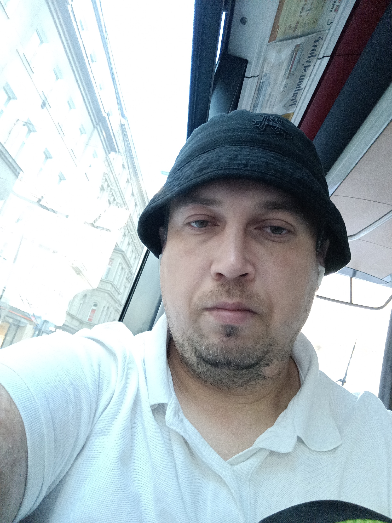 A selfie taken from the perspective of a man wearing a black bucket hat and a white polo shirt. The man has fair skin, short brown hair, and stubble on his chin. He appears to be inside a vehicle, possibly a tram or bus, as indicated by the window reflections and the interior structure visible in the background. Through the window, a building with multiple stories and windows can be seen, suggesting an urban environment. The bright daylight illuminates the scene, causing some areas to be overexposed. A newspaper or similar printed material is visible on the ceiling of the vehicle. The overall atmosphere is casual and candid.
