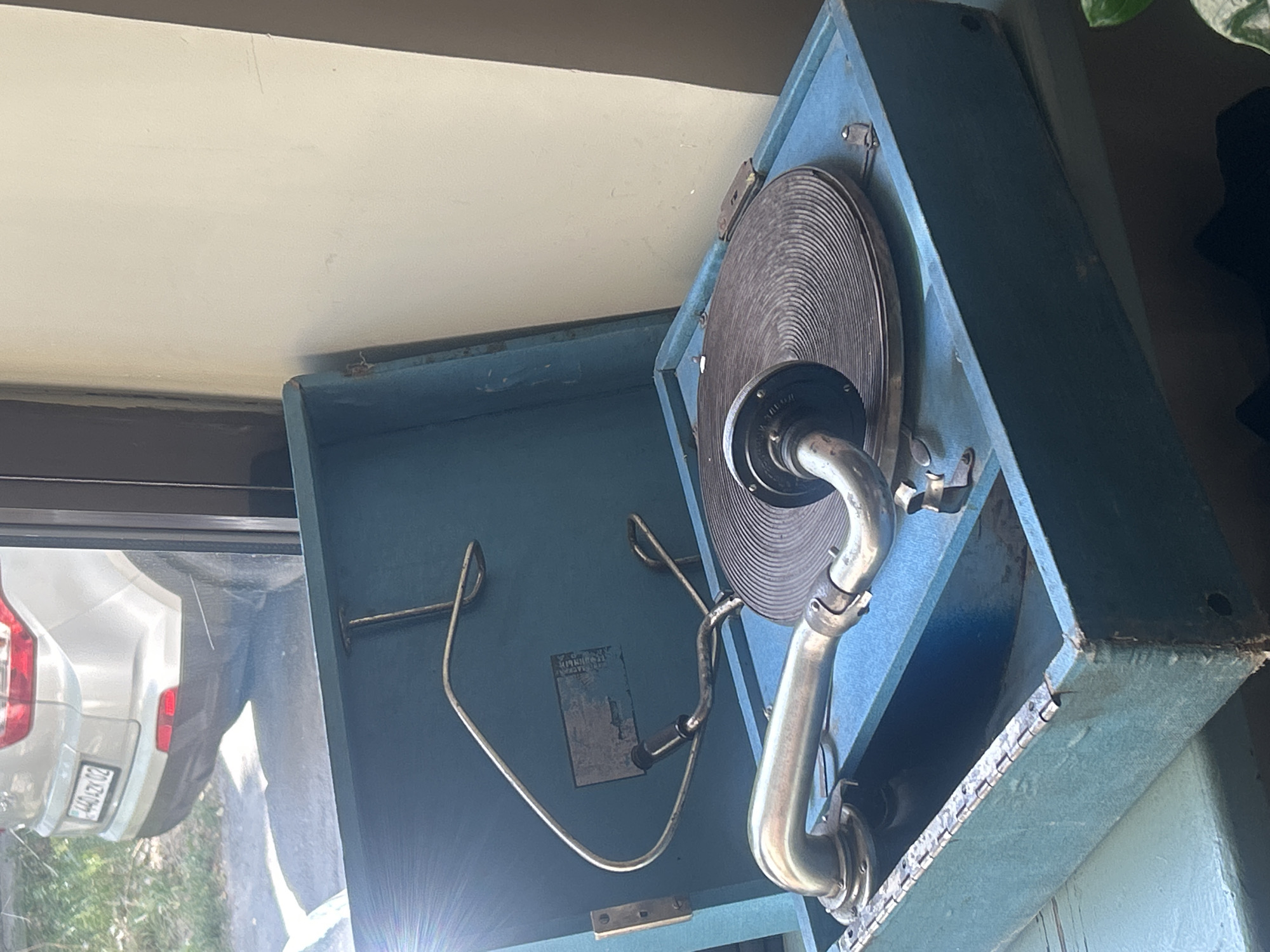 An antique blue gramophone is positioned on a teal surface, likely a windowsill or table, with its lid open. The gramophone features a circular turntable with concentric grooves and a metal tone arm ending in a horn. Behind the gramophone, a portion of a light-colored wall is visible. To the left of the gramophone, through a window, the rear of a silver-colored SUV with a license plate reading "440 AZK 02" is partially visible. The SUV is parked on an asphalt surface, with foliage in the background. This scene is located in the city of Almaty, Kazakhstan.
