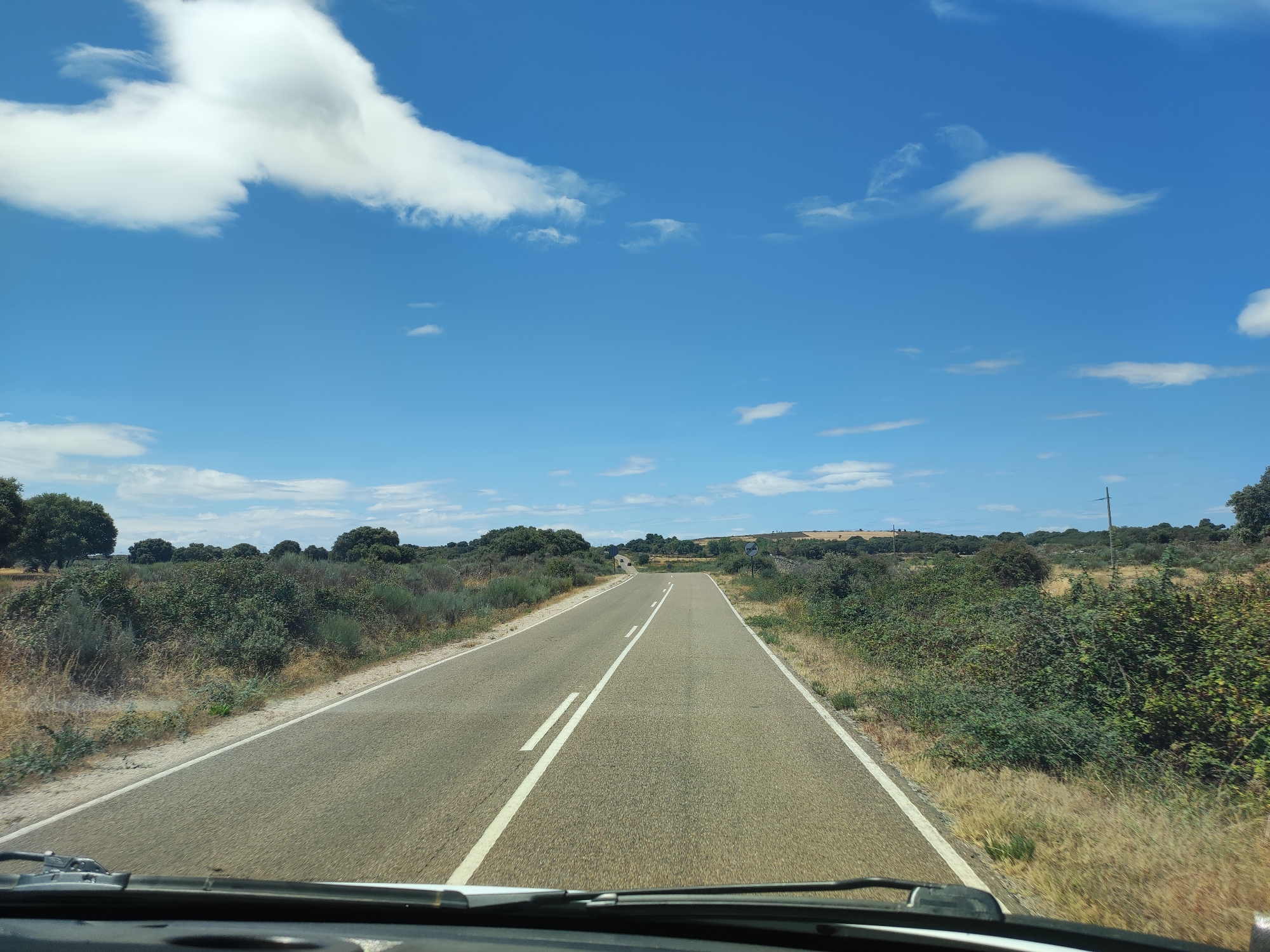 The image shows a road stretching into the distance in a rural area, with a blue sky filled with white clouds overhead. The road is paved and has white lane markings. On either side of the road, there are bushes, trees, and dry grass. In the distance, there are rolling hills. There are no people or animals visible in the image. The scene appears to be a clear, sunny day. The location is identified as a road in the city of Sobradillo, Spain, suggesting a natural, undeveloped landscape.