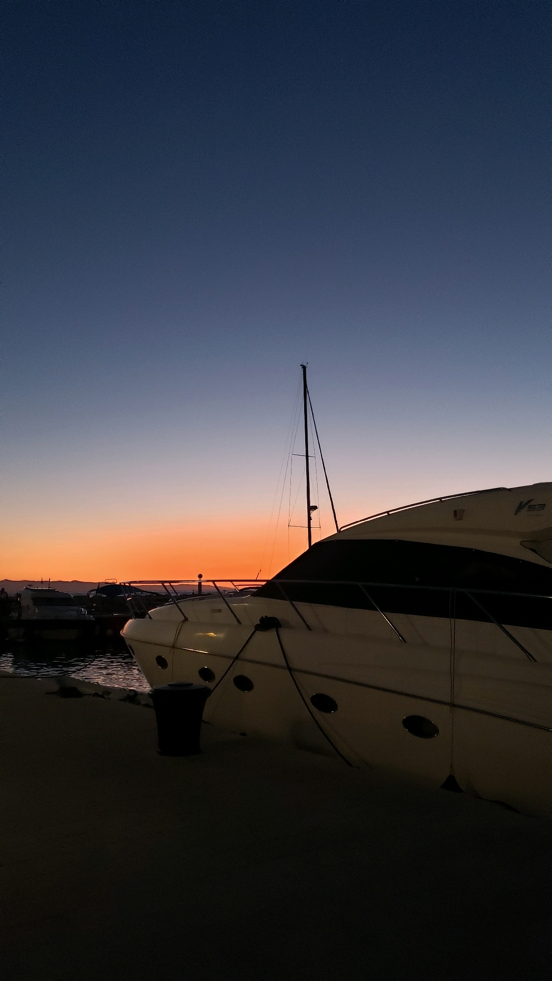 The image shows a view of a harbor in Općina Tučepi, Croatia, at dusk. A large white yacht is prominently docked in the foreground, partially covering other boats and the shoreline. The yacht has several oval windows along its hull and is secured by ropes. A tall, thin mast of a sailboat rises behind the yacht. In the background, other boats are visible, some covered, and a faint outline of hills or mountains can be seen against the horizon. The sky transitions from a deep blue at the top to a vibrant orange and pink gradient near the horizon, indicating the sunset. The ground in the foreground appears to be a paved surface, possibly a walkway or dock. A dark trash receptacle is positioned next to the yacht. The overall scene is peaceful and serene, capturing the transition from day to night in a coastal town.