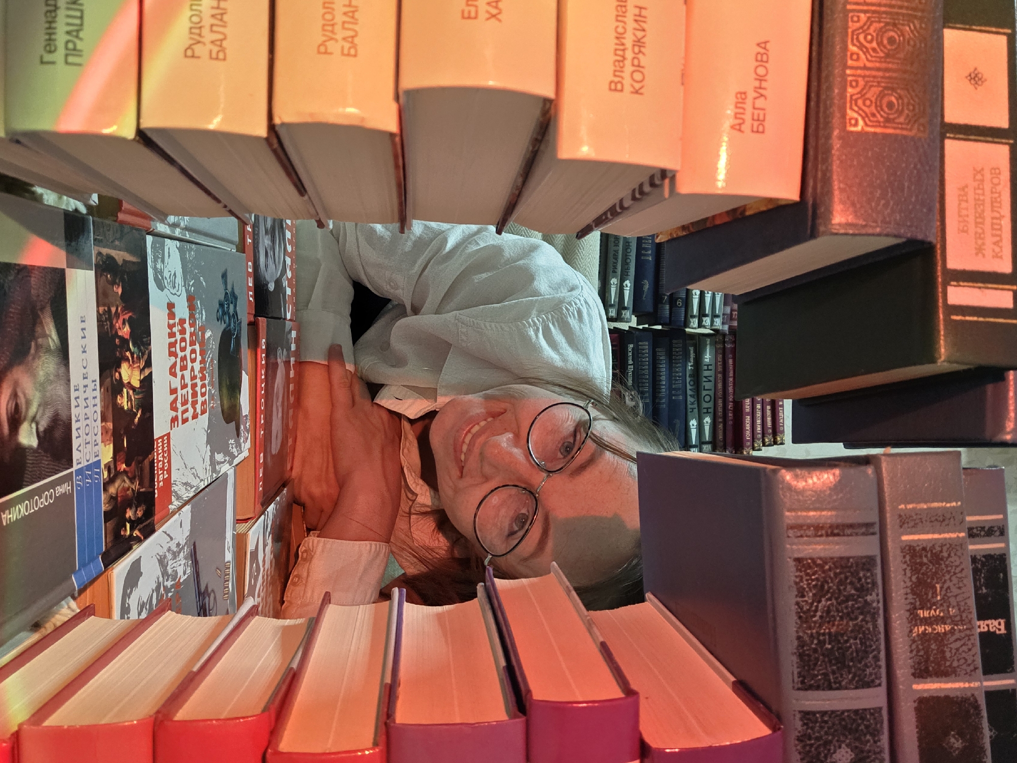 The image is taken from a low angle, looking up between two shelves of books. A person is looking down through the gap between the shelves, wearing glasses and a white shirt. Many of the book spines are in Russian, with titles like "Великие и исторические персоны" (Great Historical Figures), "Загадки России" (Riddles of Russia), and "Битва Железных Канцлеров" (Battle of Iron Chancellors). It is unknown if this image was taken in the city of Chaikovsky, Russia.