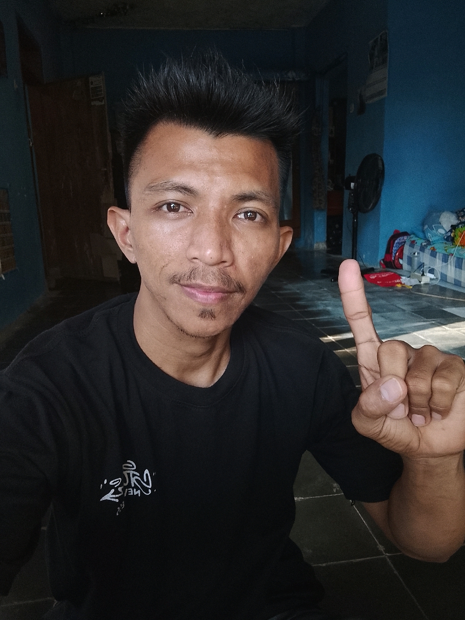 A man in a black t-shirt is taking a selfie indoors. He is looking at the camera and has his right index finger raised. The background shows a room with blue walls and a tiled floor. There is a fan in the background and some clutter on a bed. The man has short, spiky black hair and a mustache and goatee. He appears to be in his late 20s or early 30s. The image is taken in Kertahayu, Indonesia.