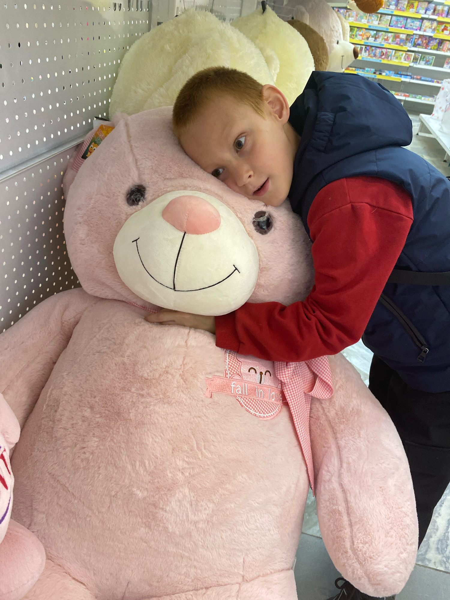 A young boy, appearing to be about 6-8 years old, with red hair and light brown eyes, is hugging a very large, plush pink teddy bear. The boy is wearing a dark blue jacket with red sleeves and black pants. His head is resting on the teddy bear's head, and he is looking towards the camera with a surprised or excited expression. The teddy bear is pink, with a large white muzzle, black button eyes, and a stitched black smile. It has a pink ribbon tied around its neck with a small tag that says "fall in love". The scene appears to be indoors, possibly in a store, with shelves of other plush toys and products visible in the background. The flooring is light-colored and tiled. The overall impression is one of affection and childlike wonder.