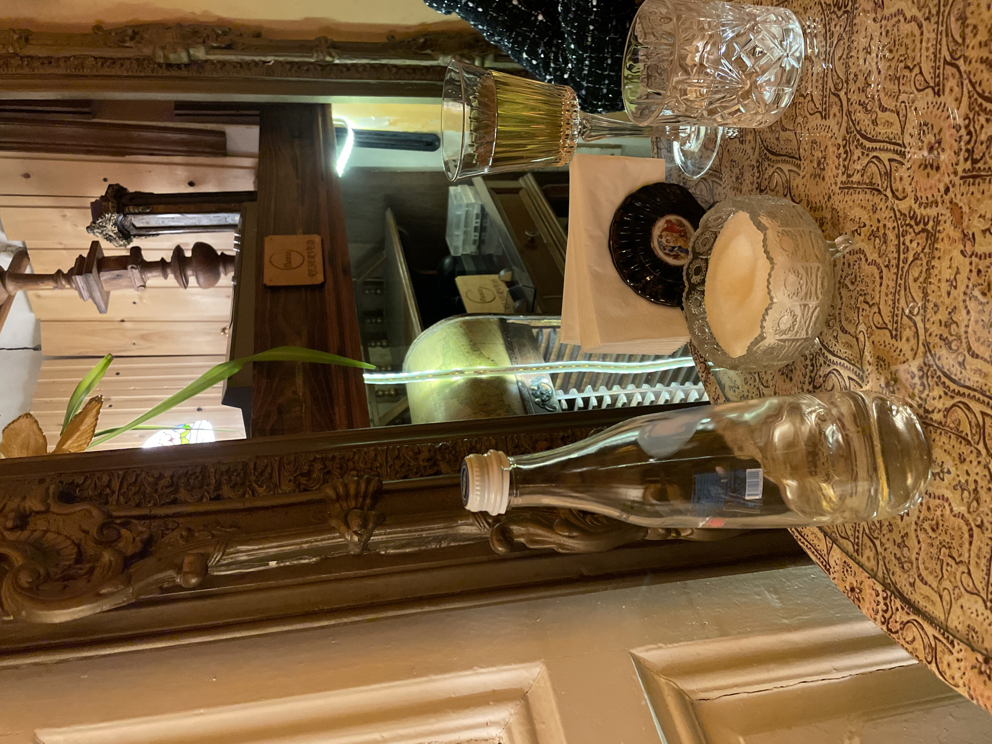 The image shows a dining table setting with reflections in a nearby mirror. On the table, covered with a patterned cloth, are two crystal glasses, a small plate with a coaster and napkins, a sugar bowl, and a bottle. The mirror reflects the table setting, along with other objects like a wooden post, plants, and architectural details. A "reserved" sign is seen in the reflection, suggesting a restaurant or bar. The lighting and decor style suggest a location in a place like Tbilisi, Georgia.