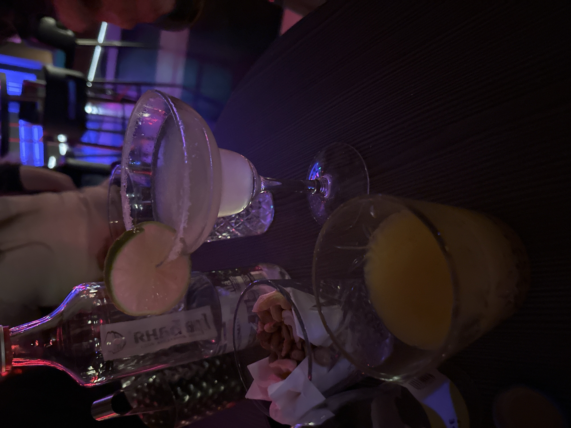 The image appears to be taken in an indoor setting, potentially a bar or restaurant, in an unknown location. It depicts a somewhat cluttered table with drinks and snacks.
**Objects:**
* **Drinks:** Two cocktail glasses, one seemingly containing a margarita with a salted rim and a lime slice, and the other filled with a yellow liquid that could be juice or another cocktail. A bottle, likely containing alcohol, is also present, with a label that is partially obscured but appears to read "RHAGAM" or similar.
* **Snacks:** A glass contains what seems to be a mixture of peanuts or other nuts, and some crumpled napkins or paper.
* **Table:** The table has a textured, dark surface.
**Environment:**
* **Lighting:** The lighting is dim with a hint of neon or colored lighting in the background, giving a somewhat nocturnal vibe.
* **Background:** The background is partially blurred, but suggests other tables and patrons may be present.
**People:**
* A partial view of a person is visible in the corner, suggesting that the image was taken from the perspective of someone seated at the table.
No city or location of Kazanlak, Bulgaria can be determined from the image.