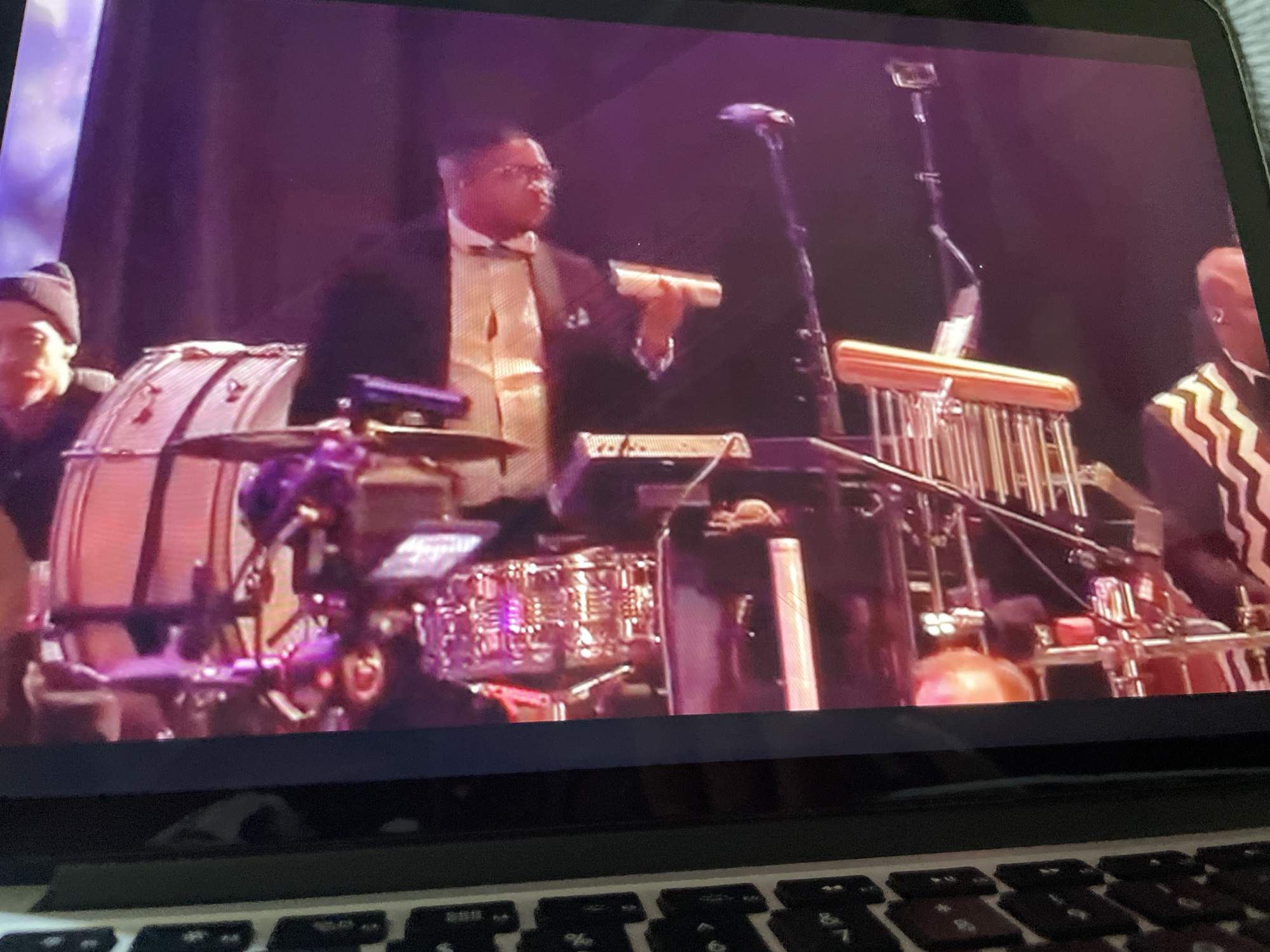 The image displays a live performance of musicians on a stage, viewed through a laptop screen which is positioned over a keyboard. In the foreground, the keyboard of a laptop is visible, with its keys partially obscured. The main subject of the image is a musician on stage, seen from the chest up, wearing glasses, a suit jacket, and a bowtie, holding a cylindrical object. To the left, another musician wearing a dark beanie and jacket is partially visible. The stage features various musical instruments, including drums, a xylophone-like instrument with metal bars, and electronic equipment. The background consists of dark purple drapes. The scene depicts an indoor concert or performance. The location in the city of Howayi, Nigeria, is not ascertainable from the provided image.