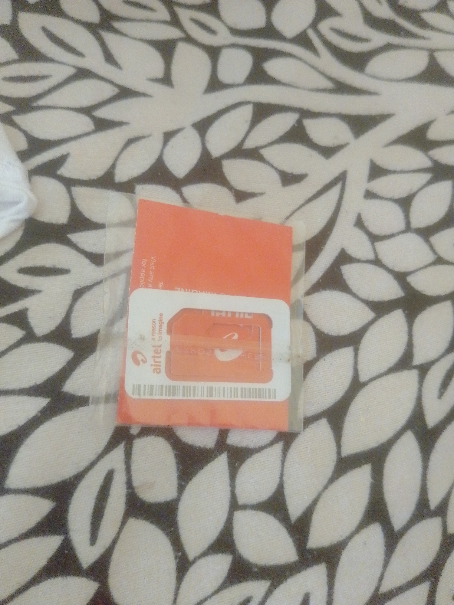 The image shows a close-up of an Airtel SIM card packet lying on a patterned fabric. The packet is predominantly orange with white lettering and graphics. A silver SIM card is visible within its cutout on the packet. The fabric has a repeating pattern of dark gray leaves on a light gray background. The lighting suggests an indoor environment, and there are no people or other objects clearly visible in the immediate vicinity. The location in Danbazau, Nigeria is not discernible from the image itself; this information is external context.
