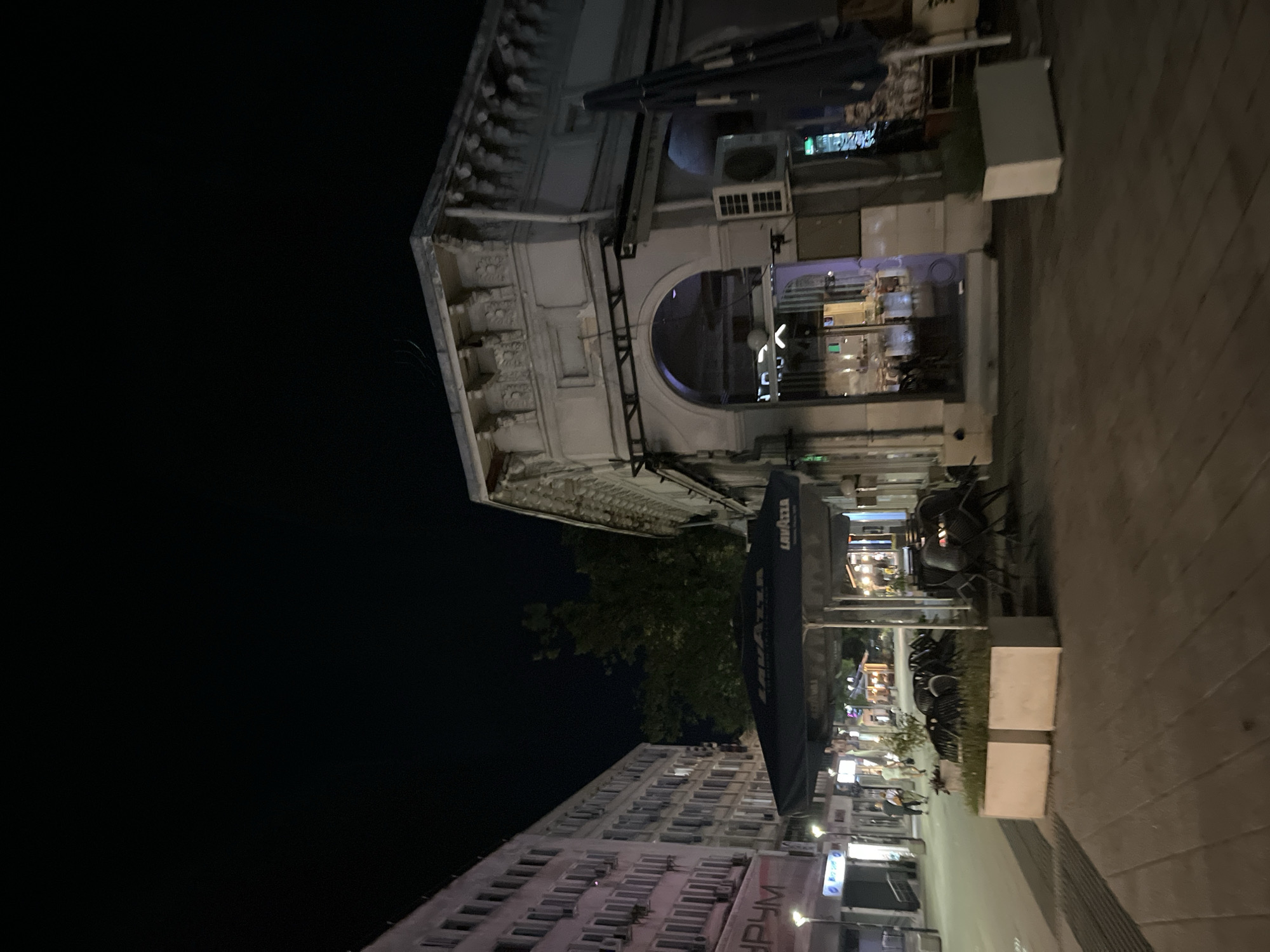 A nighttime street scene shows a city sidewalk with a large umbrella providing shade over tables and chairs. To the left, a multi-story building with many windows is illuminated, and a sign with Cyrillic text is visible. To the right, an arched doorway leads into a building with ornate architectural details. The street is paved, and planters with greenery are placed along the sidewalk. Several people are walking in the distance. The sky is dark.