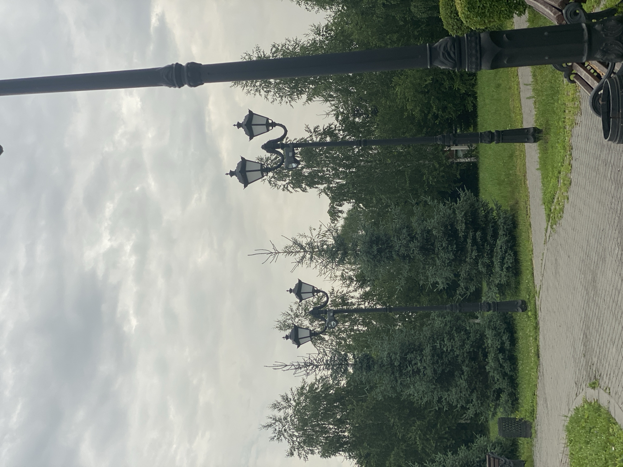 The image depicts an overcast day in a park setting, likely in Novokuznetsk, Russia, given the visual cues of mature trees and lampposts common in Russian urban parks. In the foreground, a paved walkway with a brick pattern curves to the left. To the right of the walkway, a black lamppost with decorative embellishments stands prominently. Further along the walkway and to the left, another similar lamppost with a double lantern fixture is visible. Beyond the walkway and grass, a dense line of various trees forms the backdrop, including what appear to be spruce and deciduous varieties. The sky is heavily clouded, contributing to the muted light of the scene. There are no people or animals visible in the frame. The overall impression is one of a quiet, possibly humid, late spring or summer day.