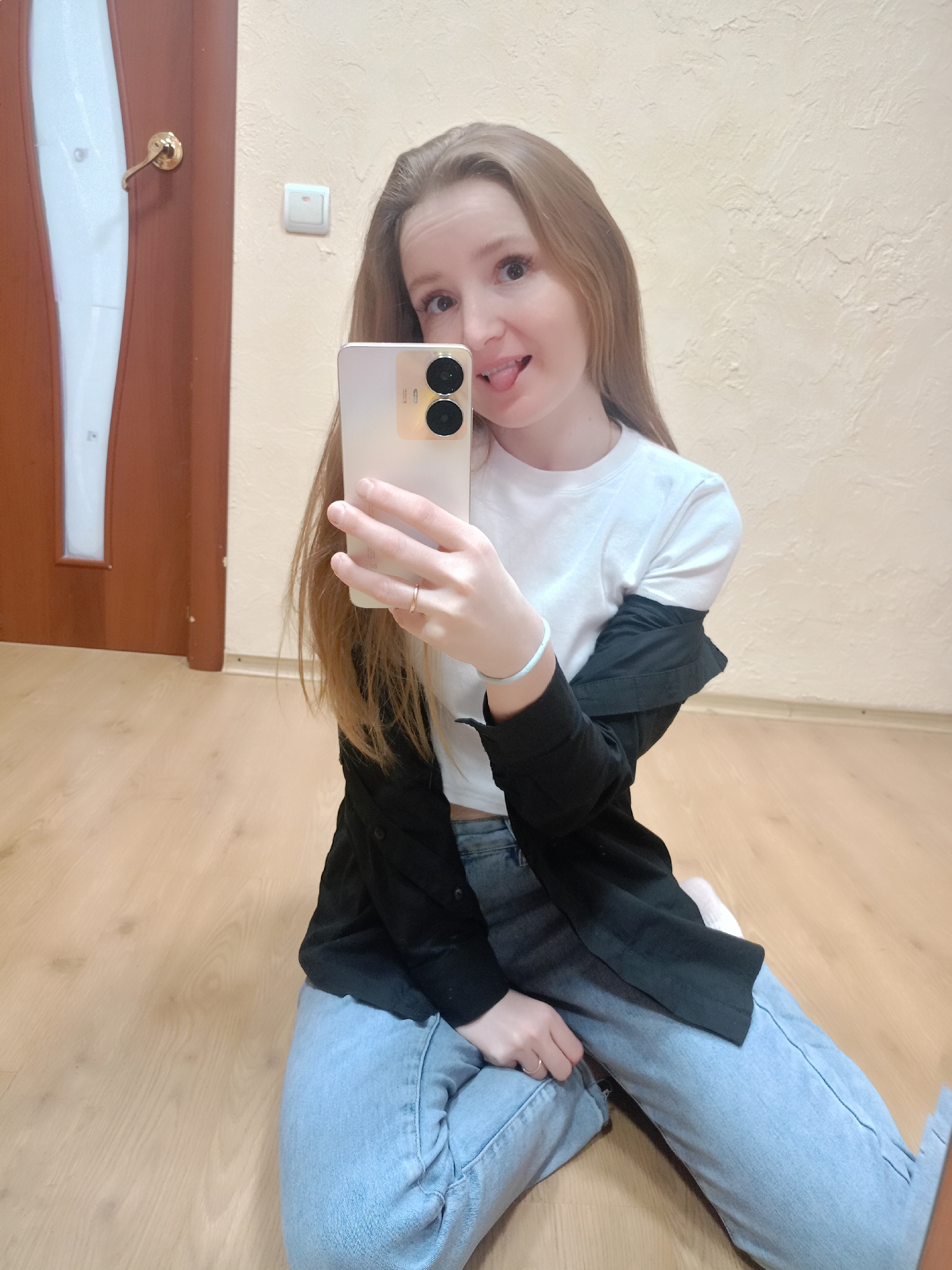 A young woman is sitting on a wooden floor, taking a selfie. She is wearing light blue jeans, a white t-shirt, and a black jacket draped over her shoulders. She is holding a gold-colored smartphone with two rear-facing cameras in her right hand, and her tongue is sticking out playfully. The background includes a door with a dark brown frame and a white panel, a light switch on the wall, and a textured light beige wall. The location appears to be indoors, potentially a room in an apartment or house. The city is Unknown, Unknown.