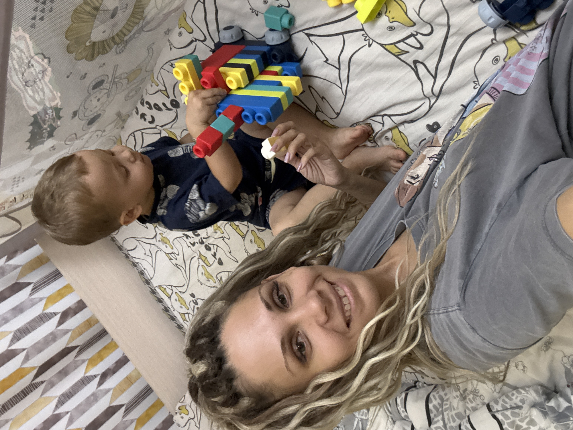 This is a close-up, eye-level, indoor, eye-level selfie taken in Rusanovo, Russia. A woman with blonde, braided hair and a grey t-shirt smiles at the camera. Beside her, a young child in a dark blue t-shirt plays with colorful building blocks. The background is a bed with a patterned duvet depicting cartoon animals. The overall scene is relaxed and playful, suggesting a casual moment between a mother and child.